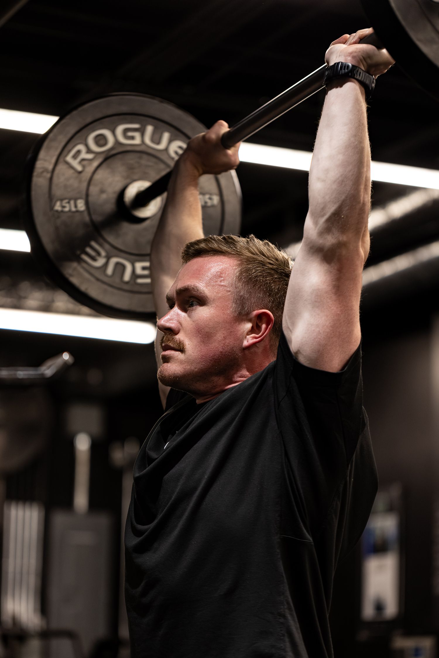 Focused male athlete pressing Rogue barbell overhead in dark gym, wearing black t-shirt with visible effort.