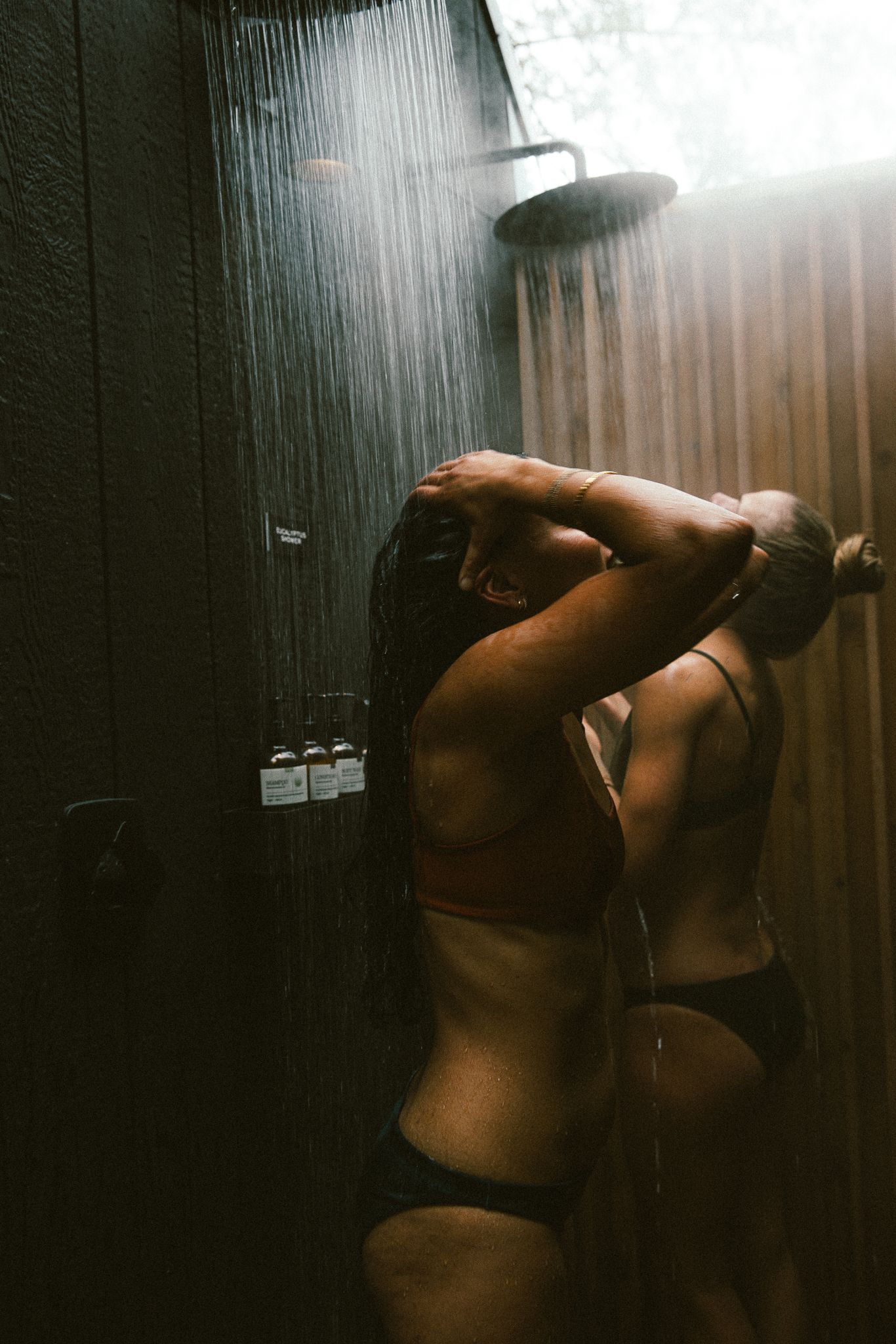 Woman in bikini showering under rainfall showerhead in a dark wooden outdoor shower with dramatic lighting.