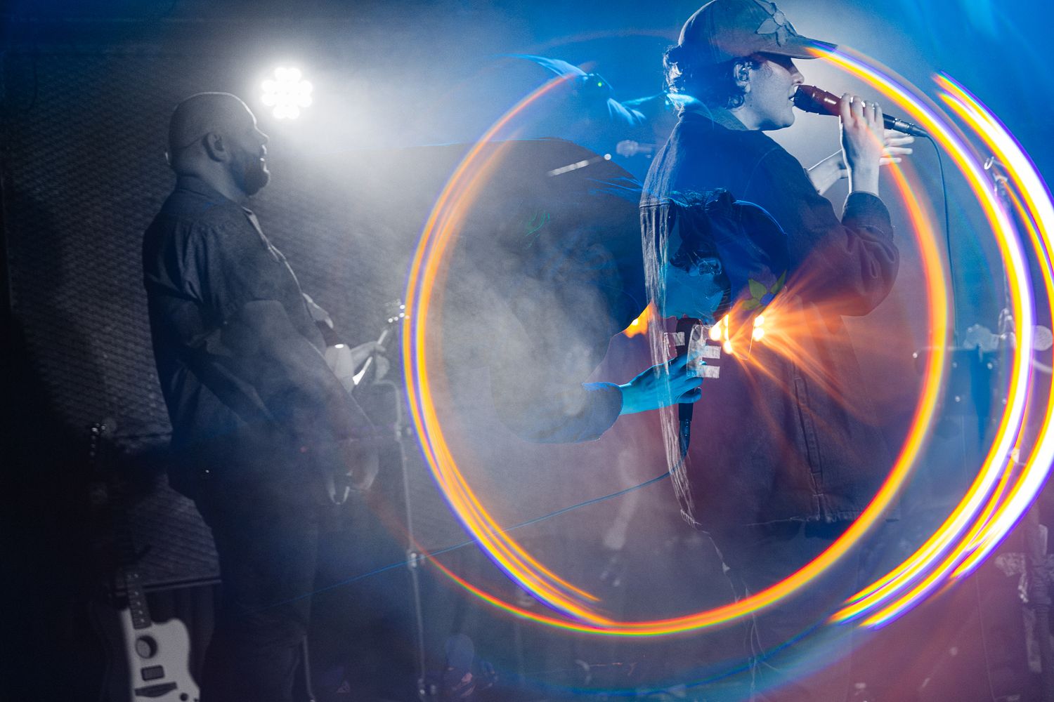 Two musicians perform on a smoky stage with dramatic orange circular light trails swirling around them.