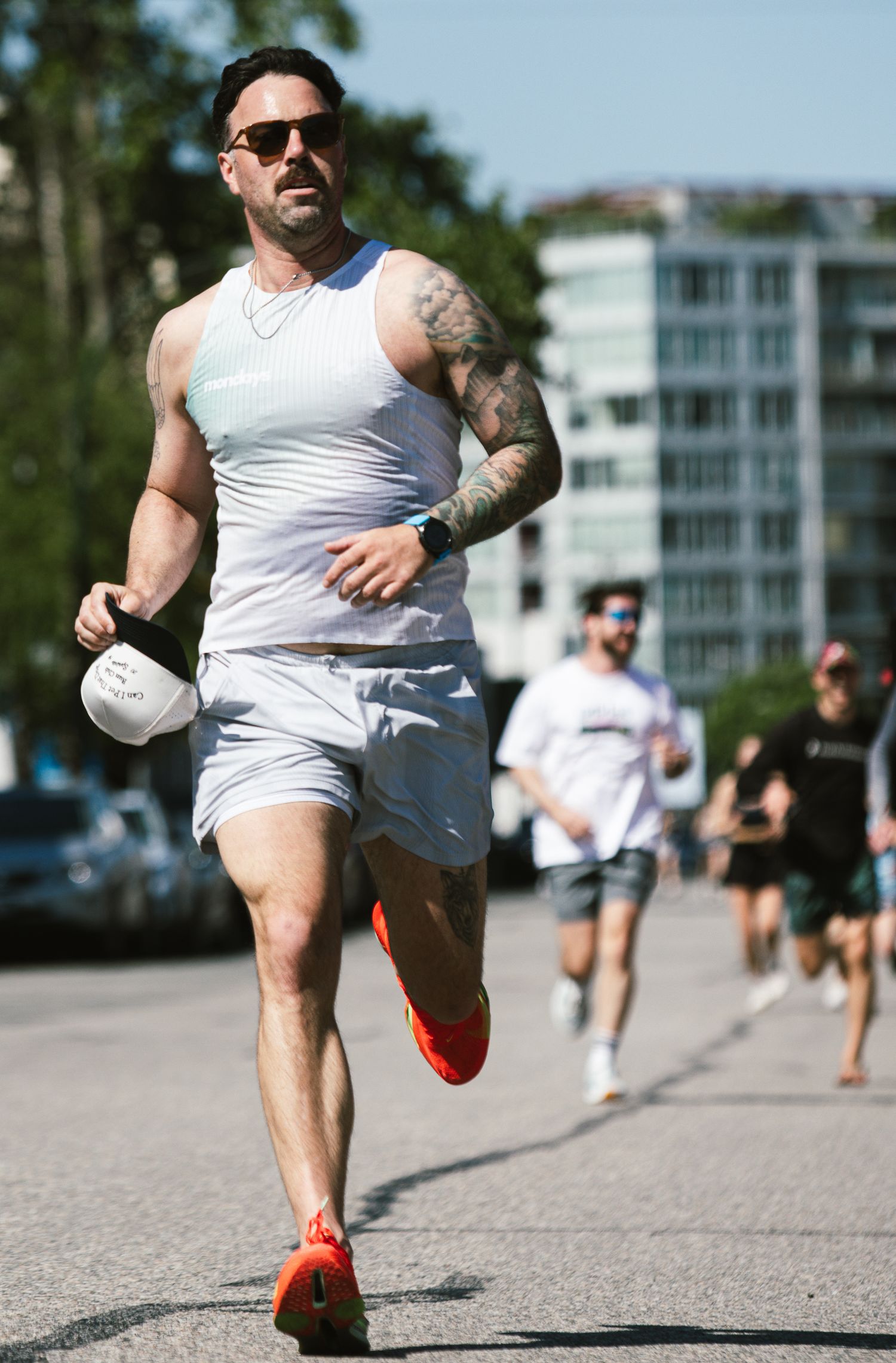 Tattooed male runner in white tank and shorts sprints down sunny urban street wearing orange shoes.