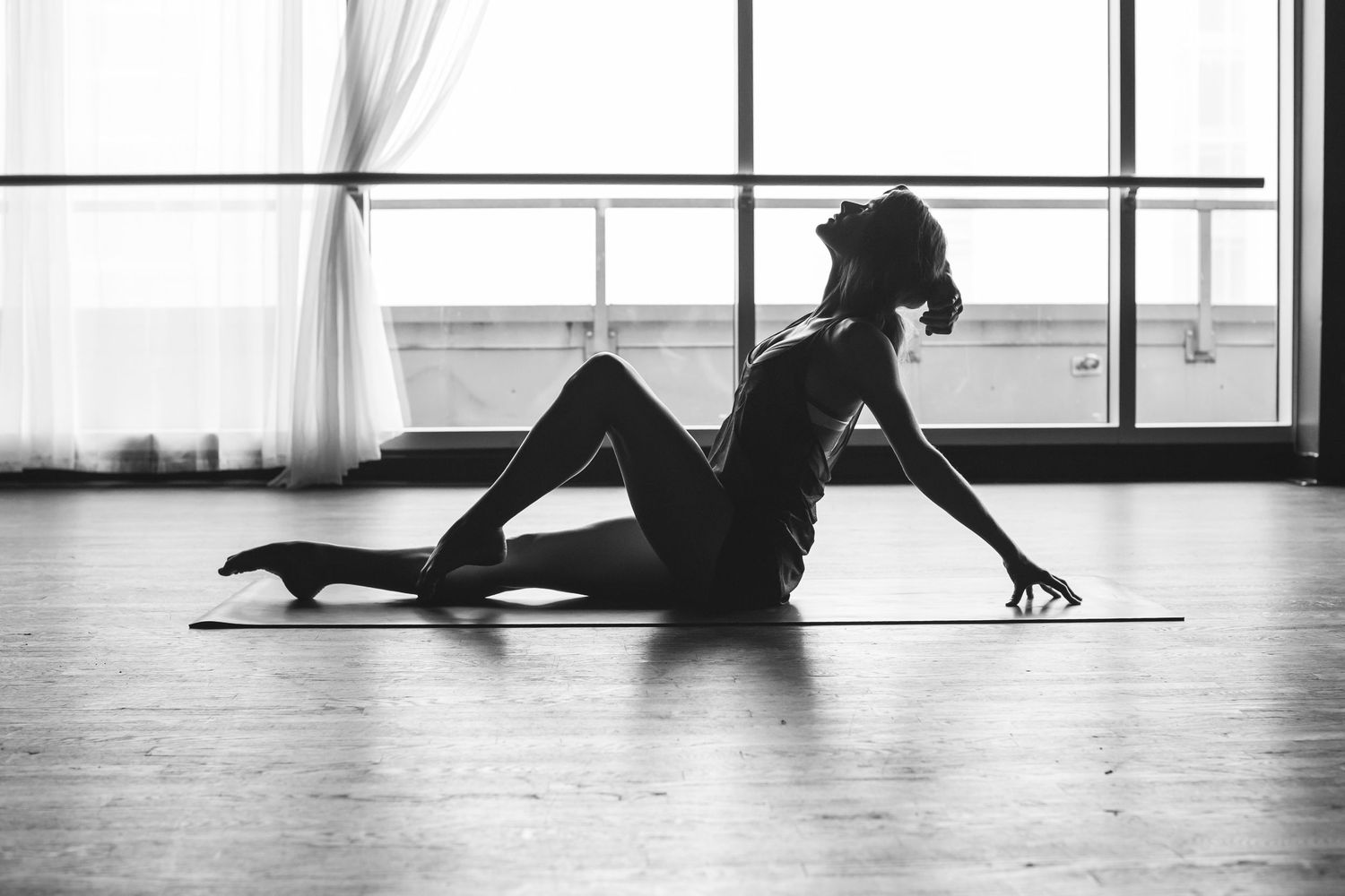 Black and white photo of a woman in a yoga pose on a mat in a bright studio with large windows.