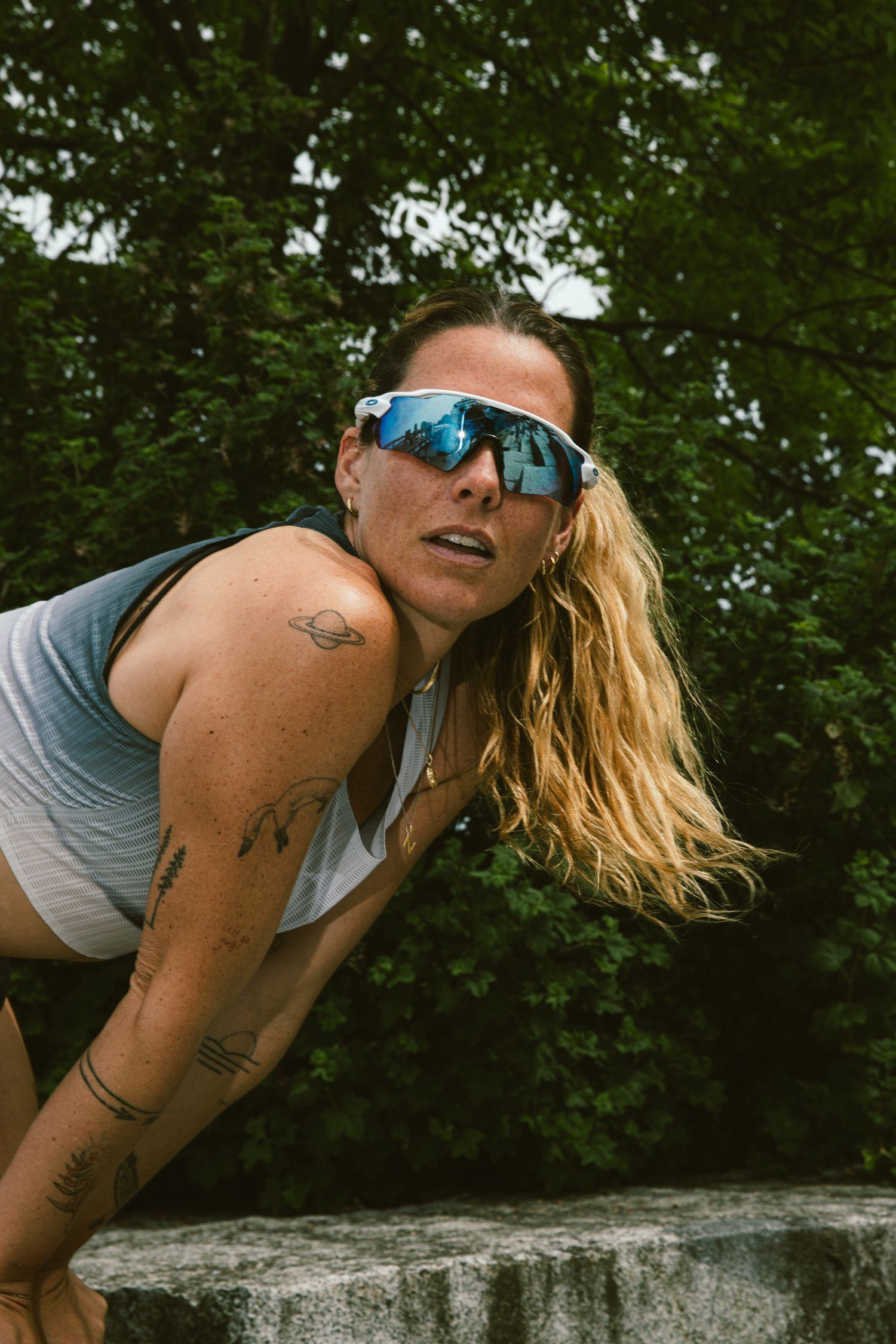 Athletic tattooed woman in sport sunglasses leaning forward outdoors near greenery.