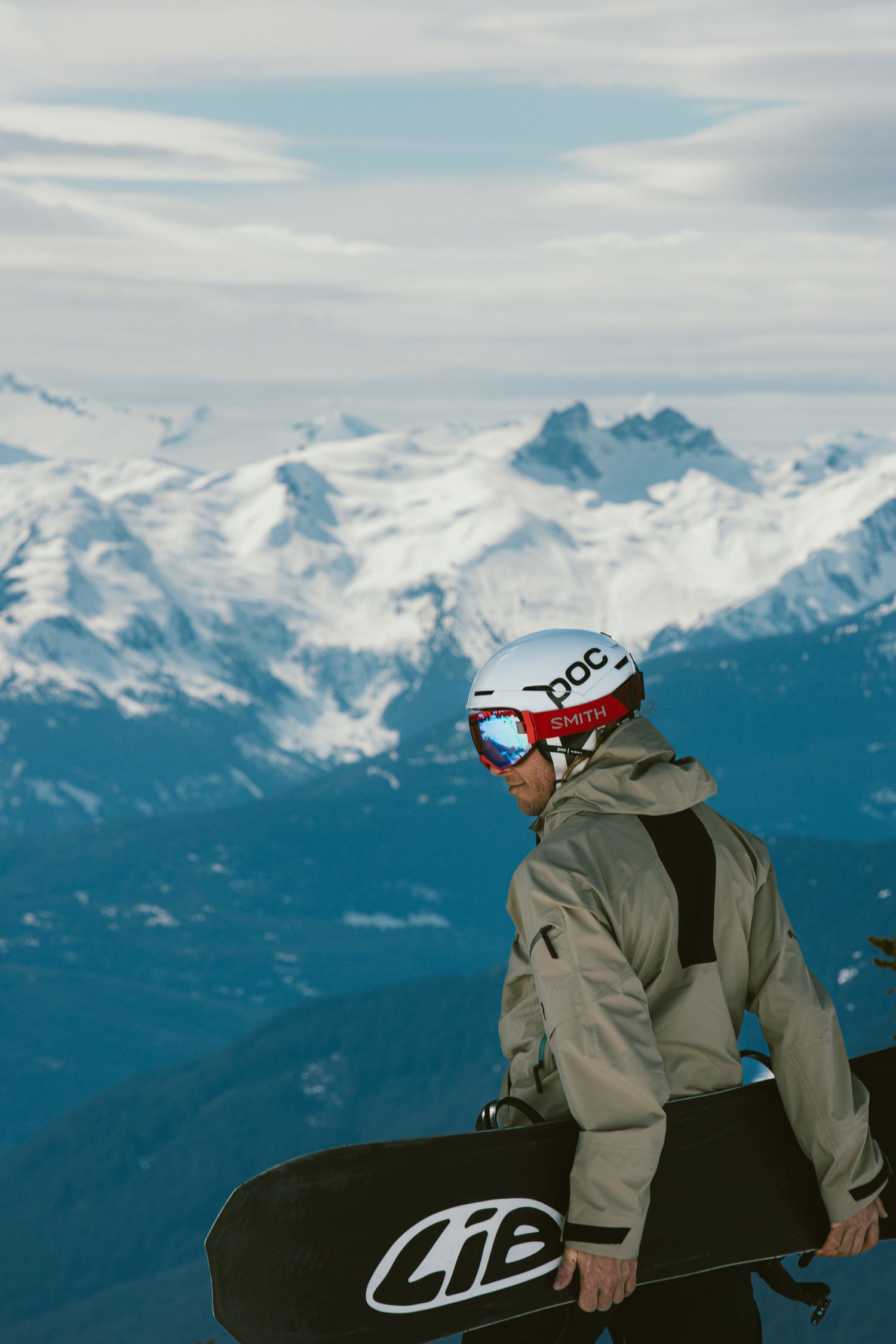 Snowboarder holding Lib Tech board overlooking snow-capped mountain range wearing POC helmet.