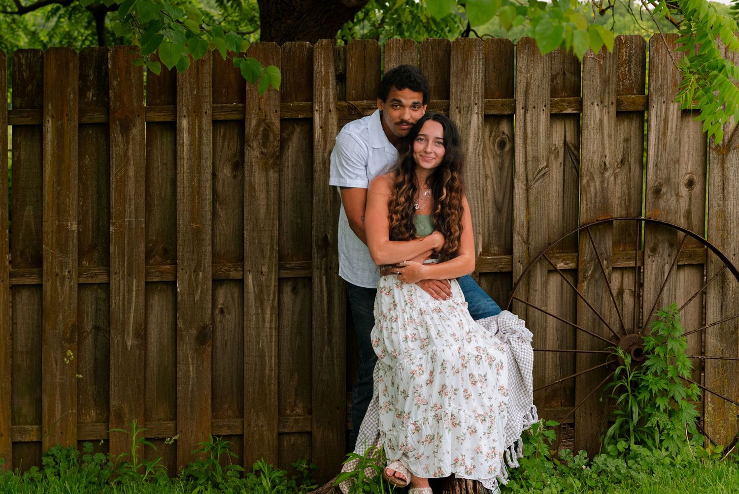 A couple posing affectionately in front of a rustic wooden fence with a decorative wagon wheel.