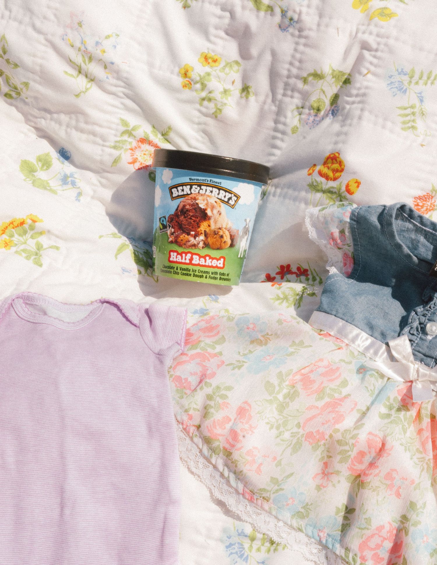 Ben & Jerry's Half Baked ice cream surrounded by baby clothes on floral quilt.