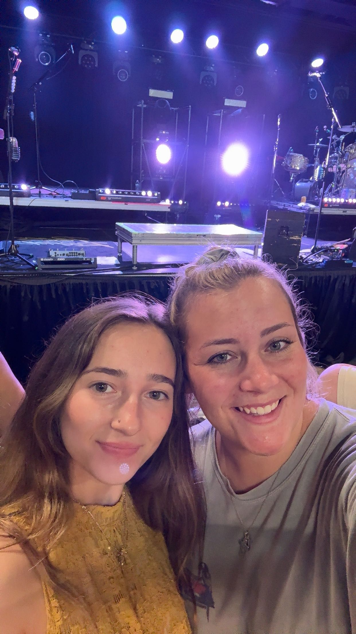 Two women smiling for a selfie at a concert venue with colorful stage lights and instruments in the background.