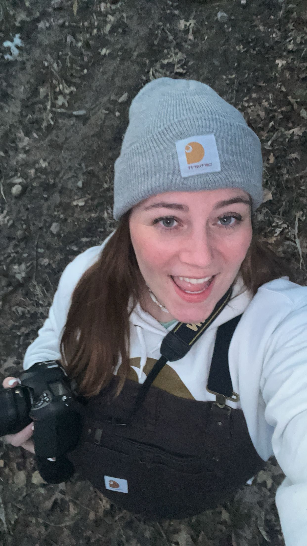 Smiling woman wearing a gray Carhartt beanie and overalls holds a DSLR camera outdoors in a wooded area.