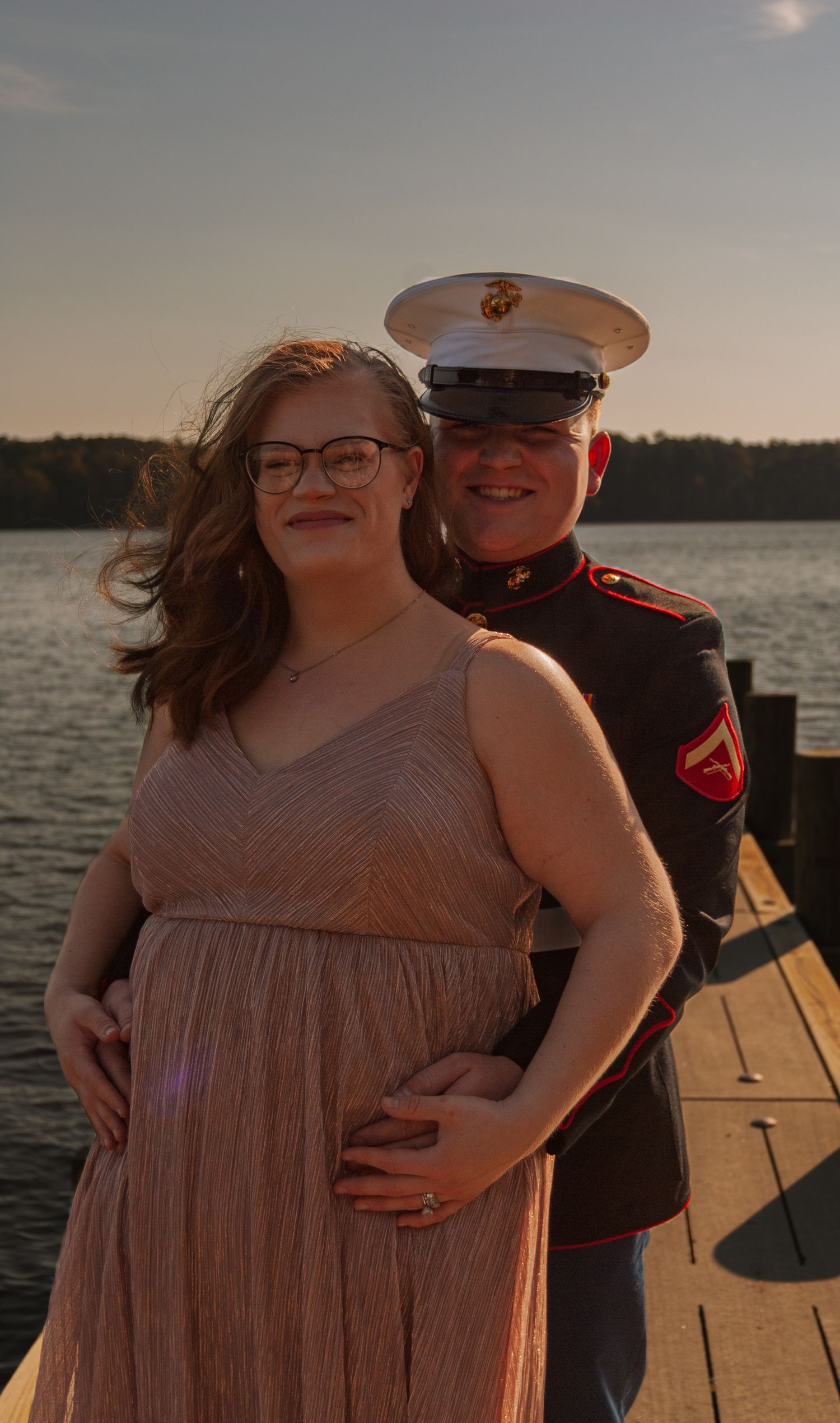 A Marine in dress uniform embraces a woman in a pink gown on a lakeside dock at sunset.