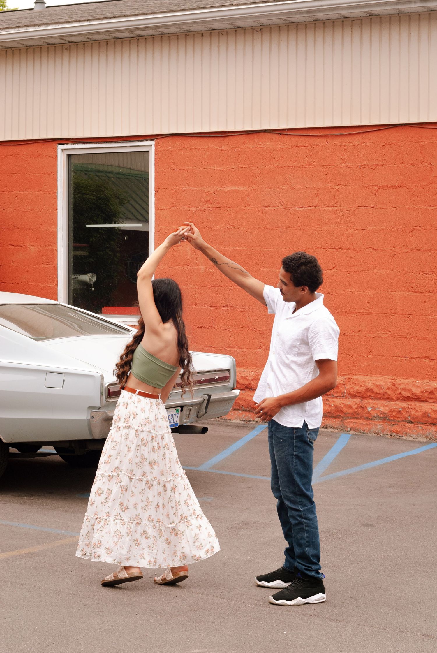 Couple dancing outdoors in a parking lot with a classic white car and orange brick wall behind them.