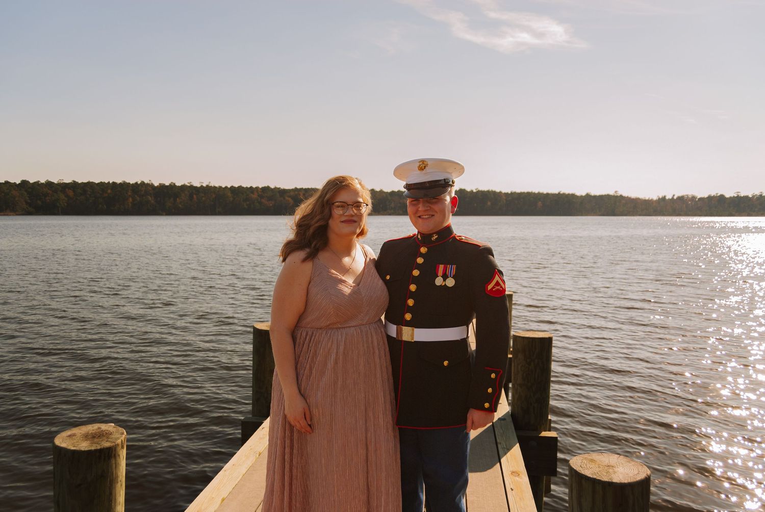 A woman in pink gown and a US Marine in dress blues pose together on a sunny dock.