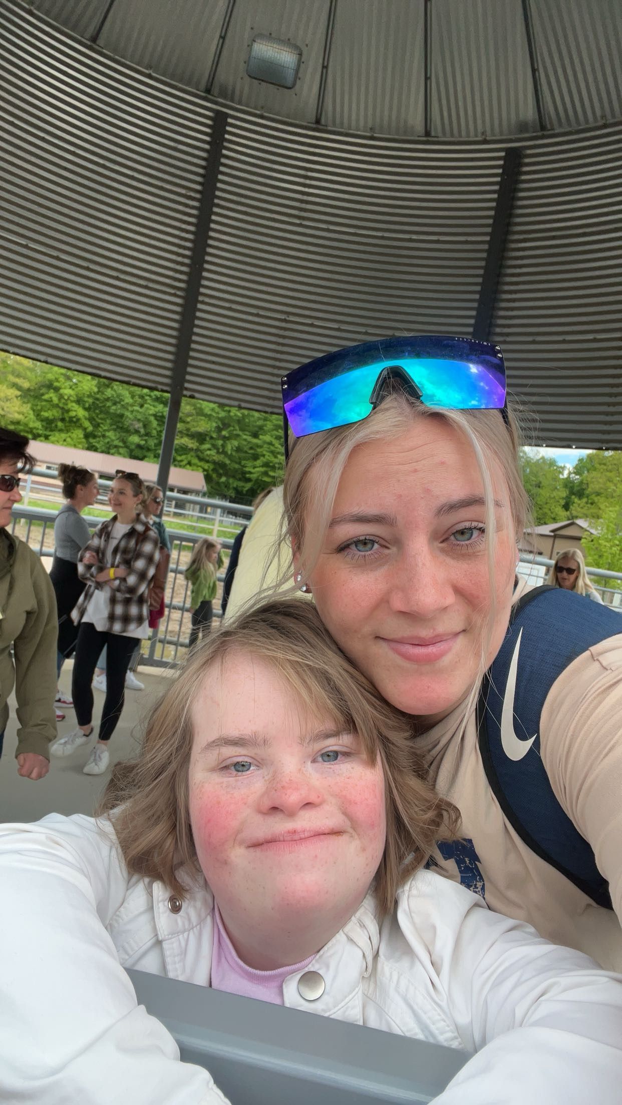 Two smiling women take a selfie under a pavilion, one wearing blue mirrored sunglasses on her head, outdoors.