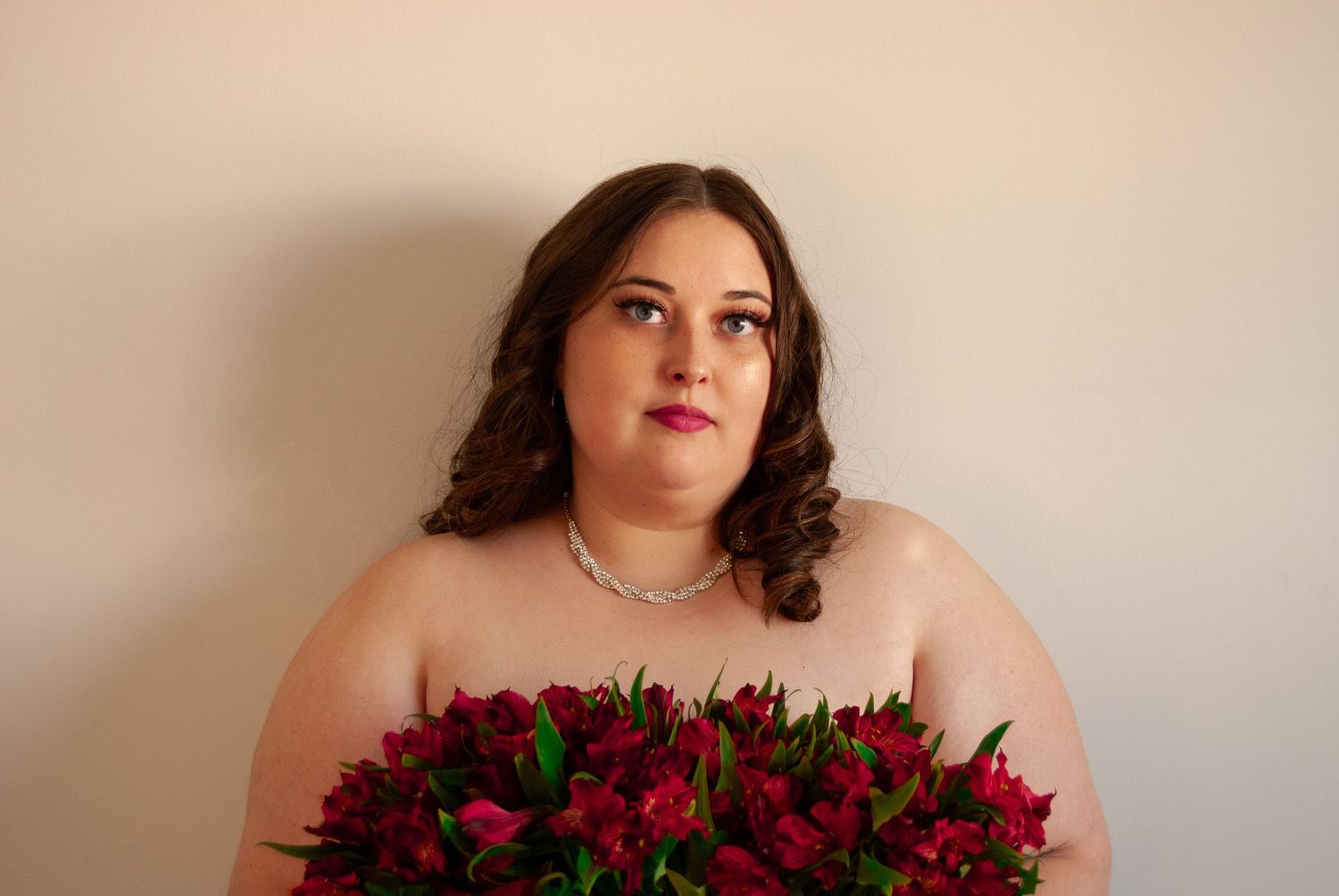 Plus-size woman with pearl necklace holds red flowers, gazing upward against a light background.
