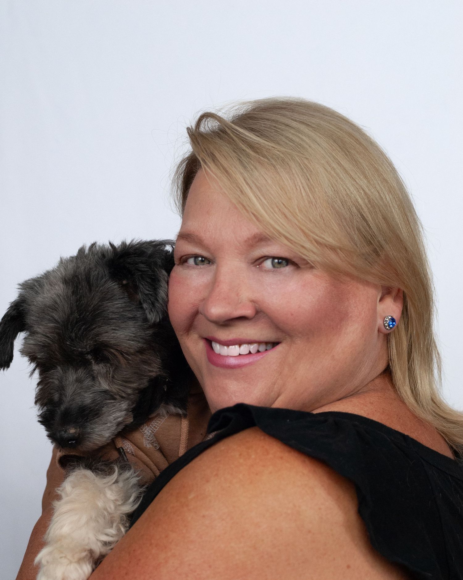 A smiling blonde woman in a black top holds an adorable small gray and white puppy.