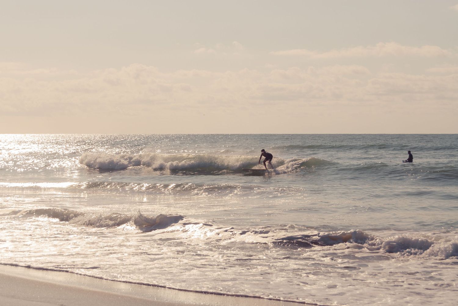 Two surfers riding small waves on a bright, hazy beach morning.
