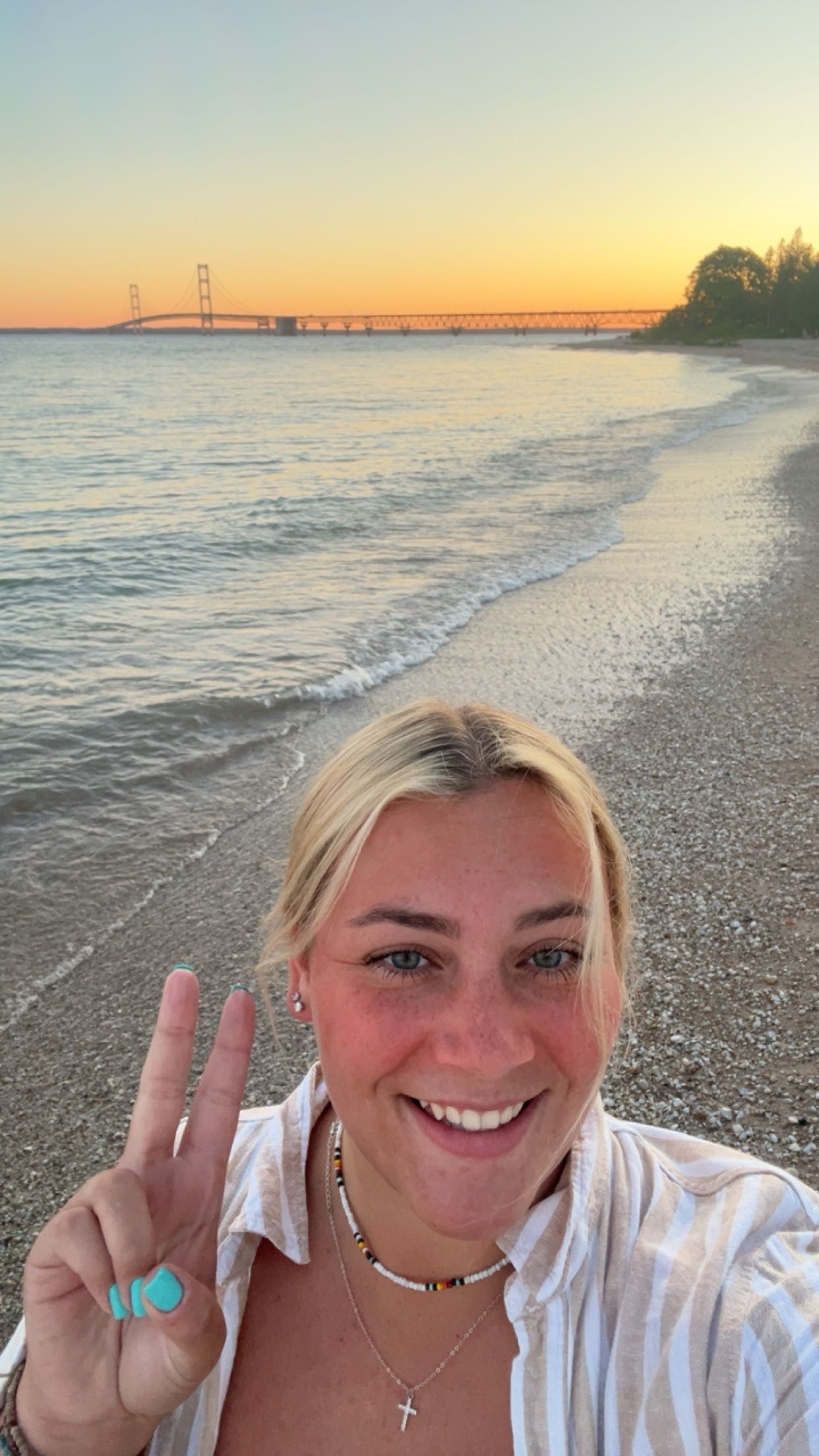 Smiling blonde woman taking a selfie on a sandy beach at sunset with a long bridge visible in the background.