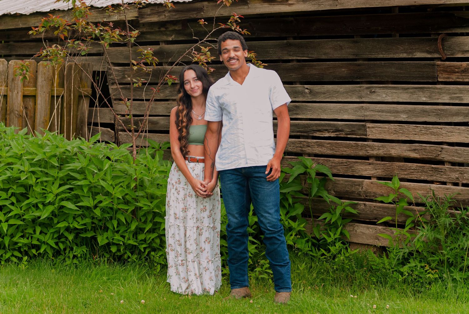 A young couple smiling and posing together in front of a rustic log building on green grass.