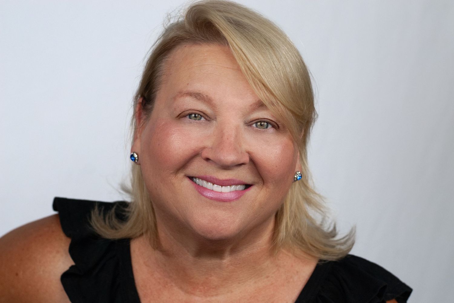 A cheerful blonde woman wearing blue earrings and a black ruffled top in a professional portrait.