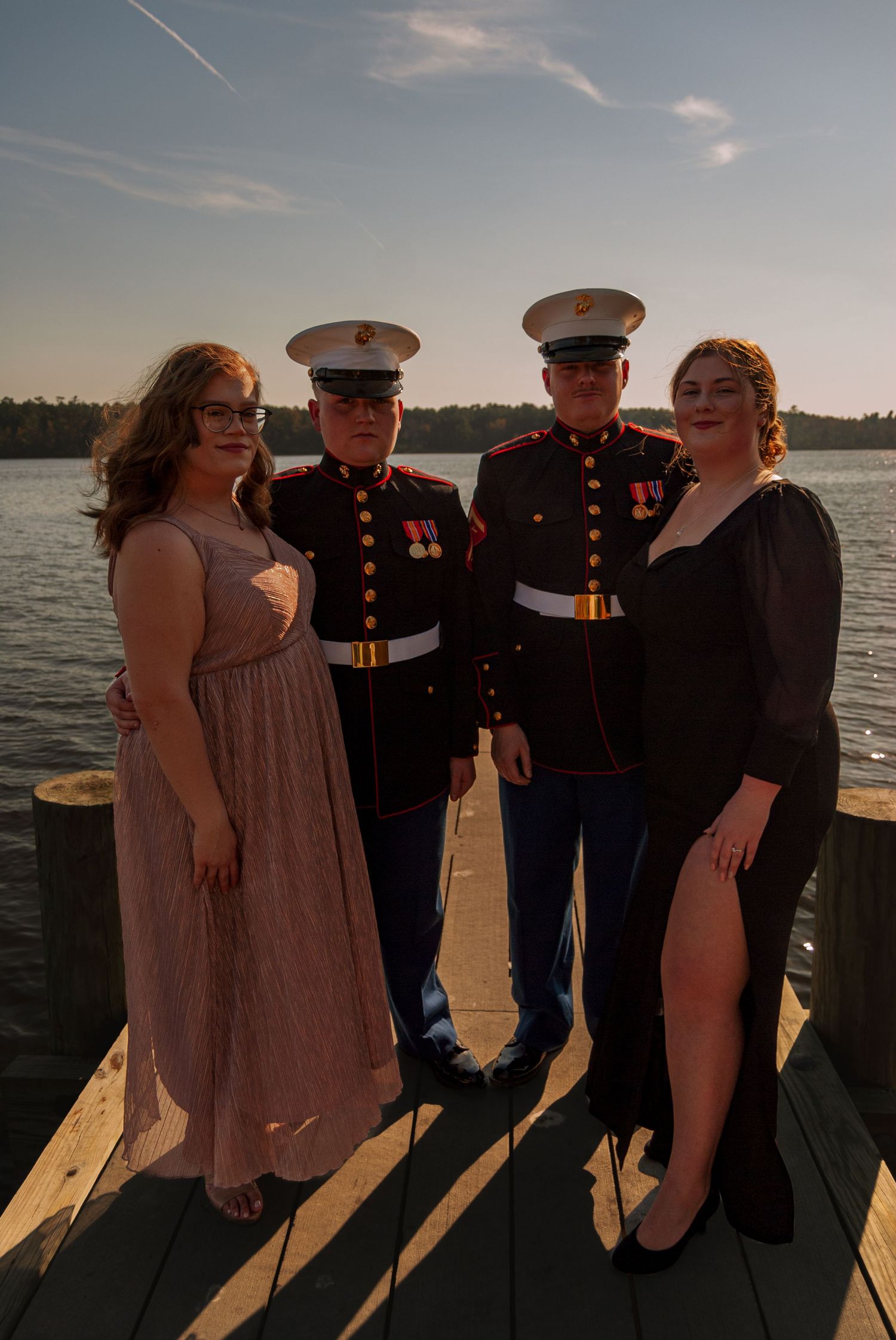 Two Marines in dress uniforms pose with two women in formal gowns on a waterfront dock at sunset.