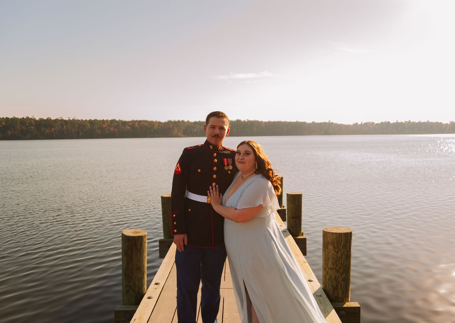 A couple poses on a lakeside dock, man in Marine dress blues, woman in white dress.