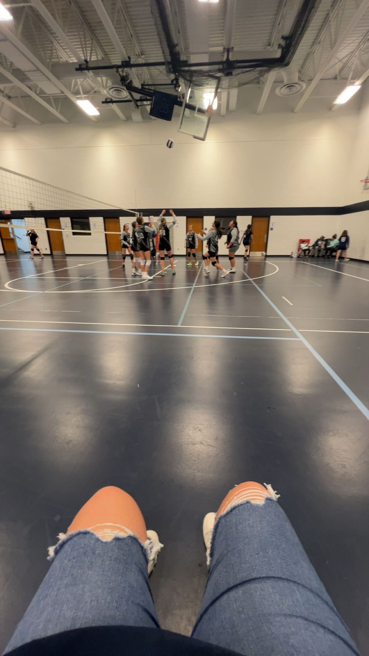 Person watching a youth volleyball game in a school gymnasium, with players gathered near the net on a dark hardwood court.