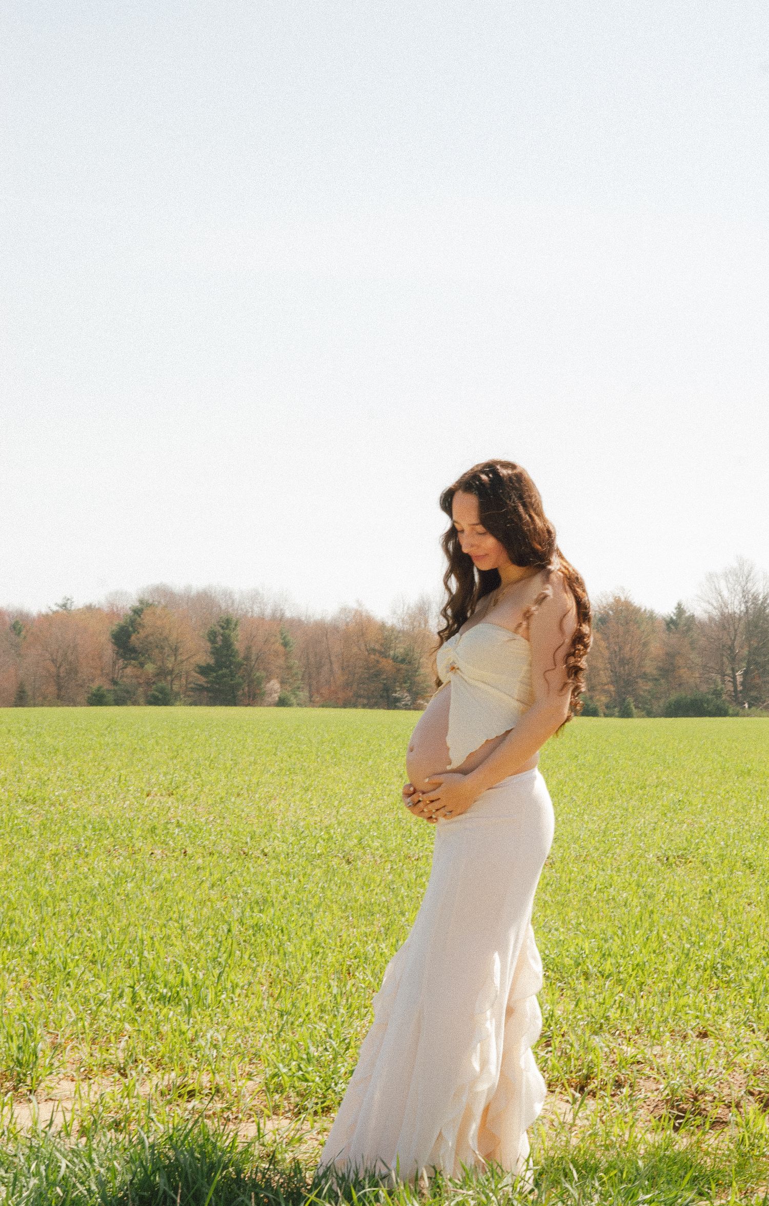 Expectant mother in white lace gown gazes down at baby bump in open field.