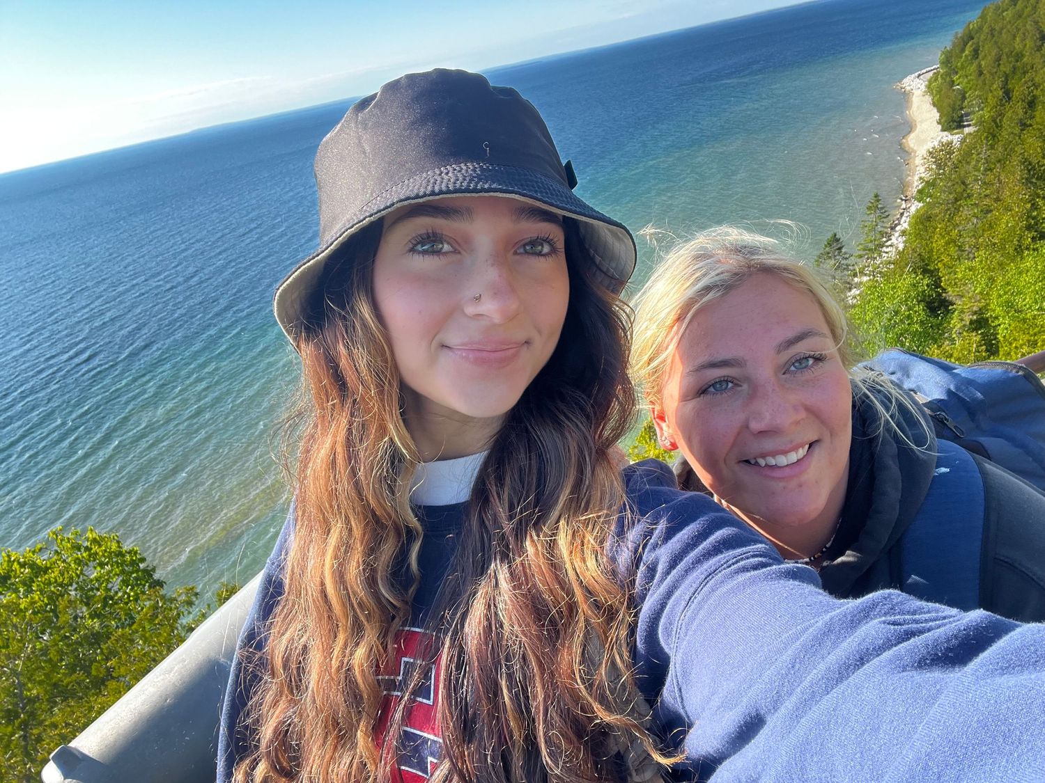 Two women taking a selfie outdoors with a scenic ocean and forested cliff view in the background.