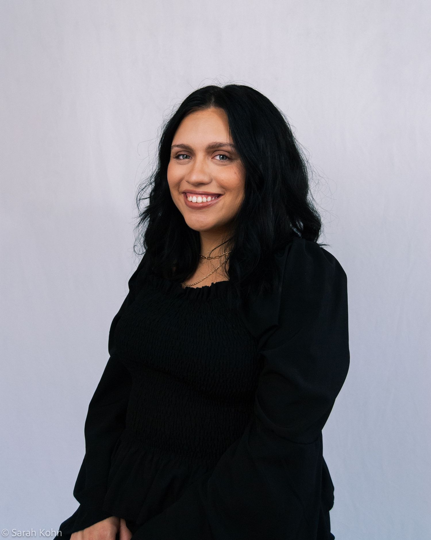 Smiling woman with long black wavy hair wearing a black top, posing against a light gray background.