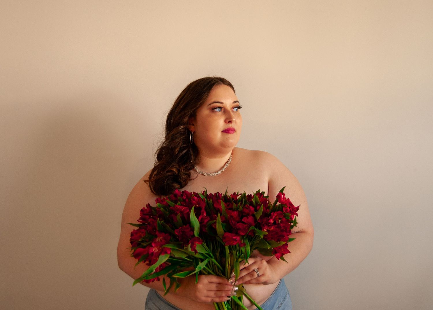 Plus-size woman in strapless top holds vibrant red alstroemeria bouquet against a neutral backdrop.