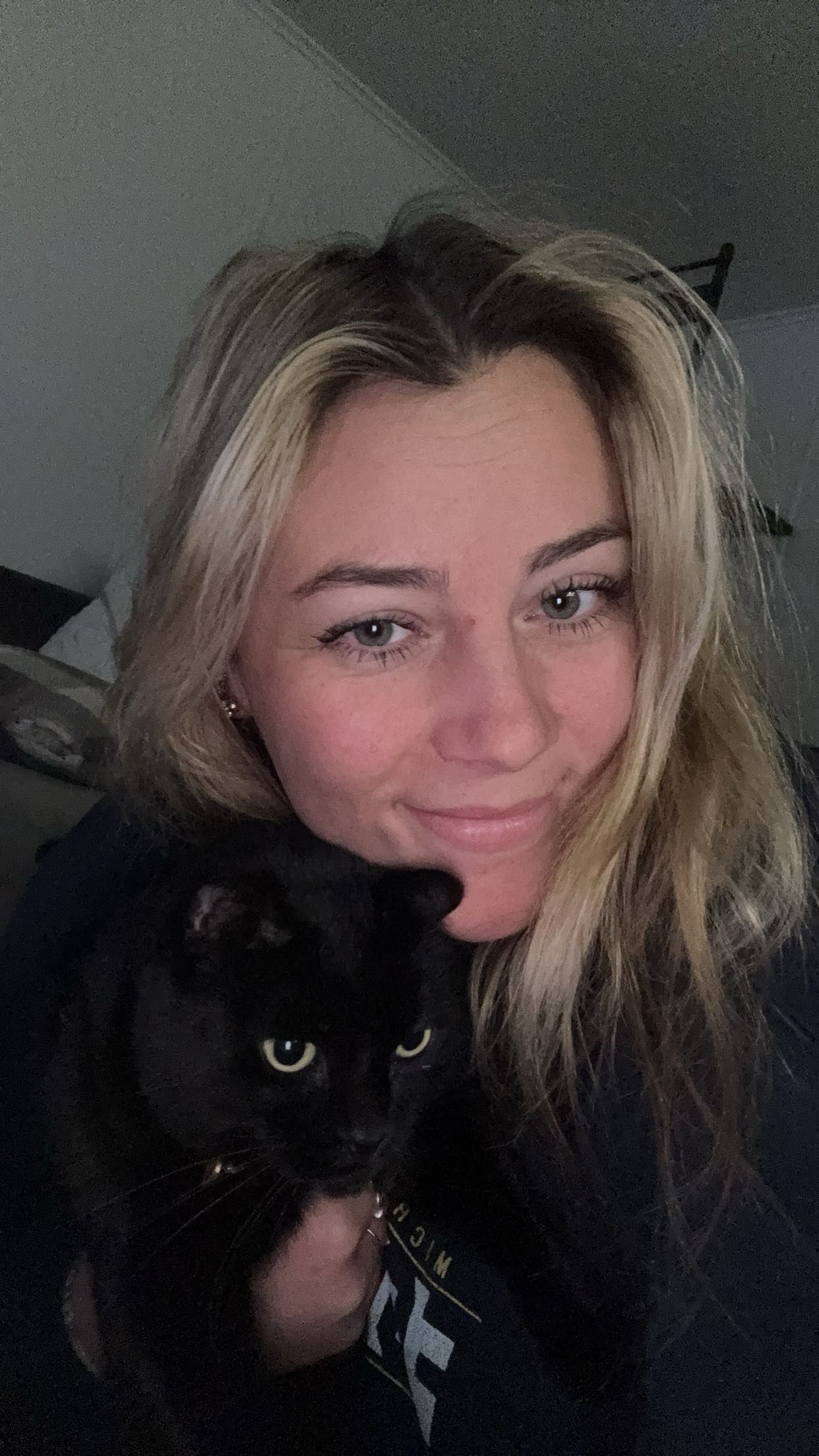Smiling blonde woman holds a black cat close to her face indoors for a casual selfie portrait.