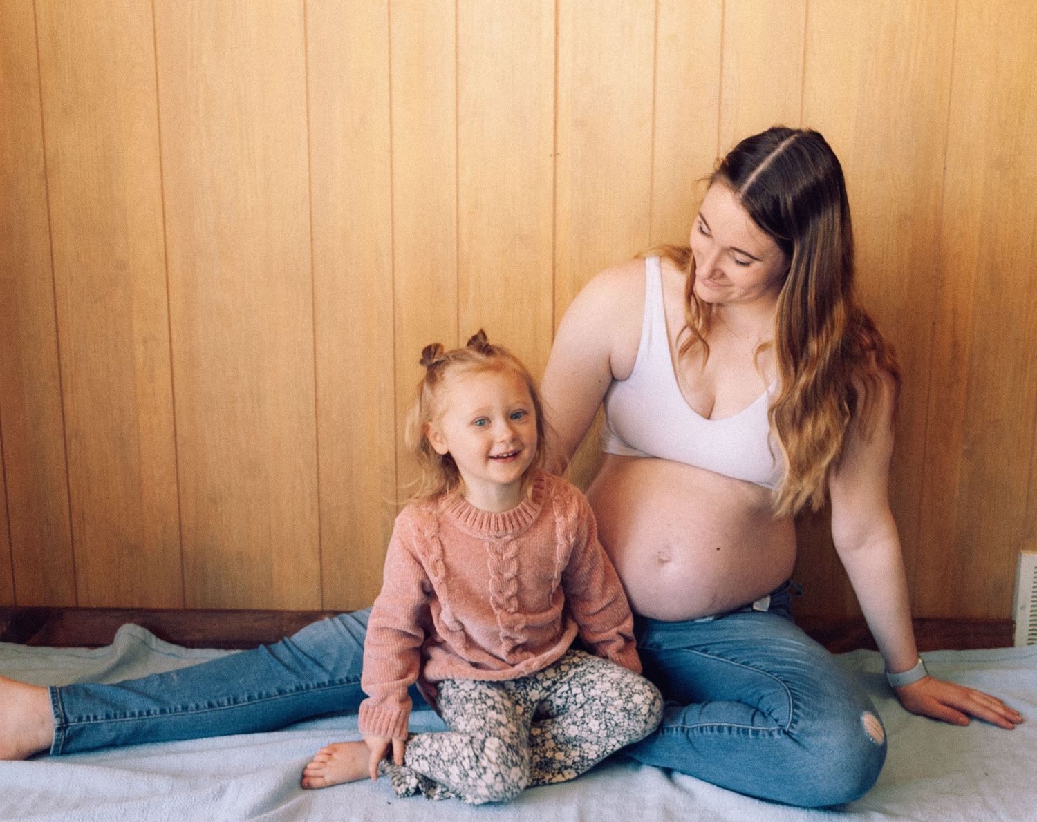 Pregnant woman in white bra and jeans sitting with laughing toddler girl indoors.