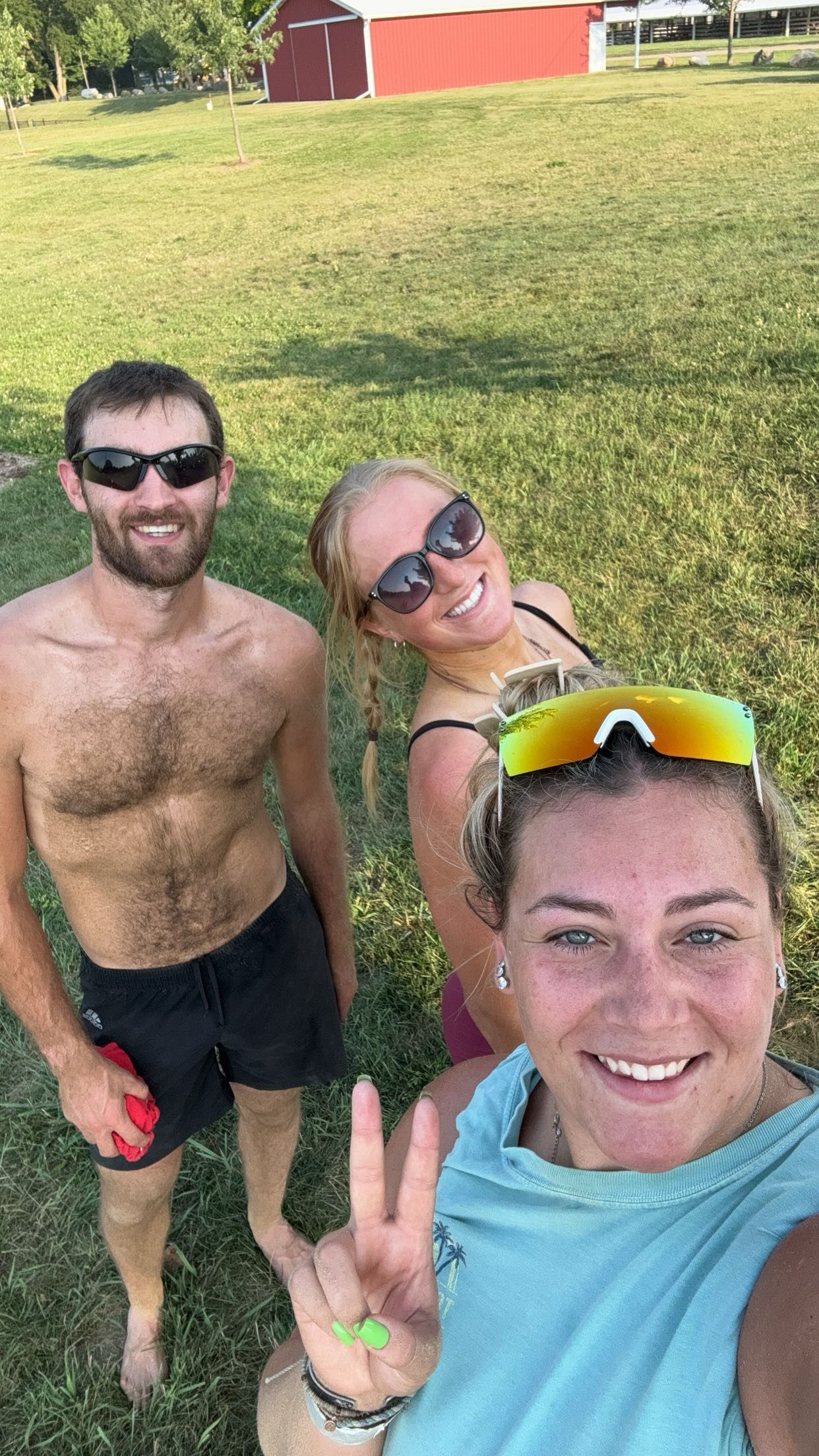 Three friends smiling for a selfie outdoors on a sunny day near a red barn and green field.