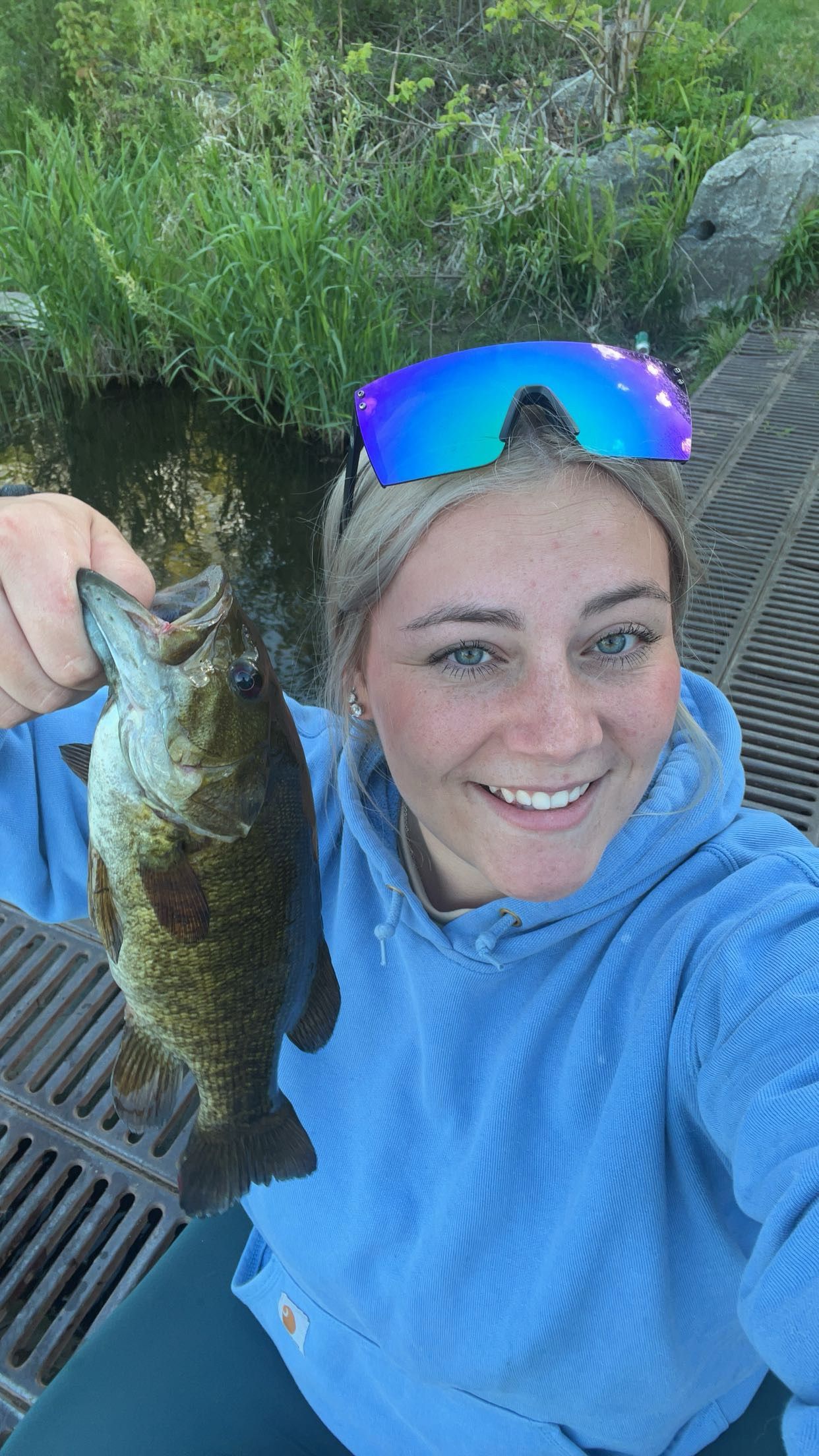 Smiling blonde woman holding a largemouth bass while fishing, wearing a blue hoodie and colorful sport sunglasses outdoors.