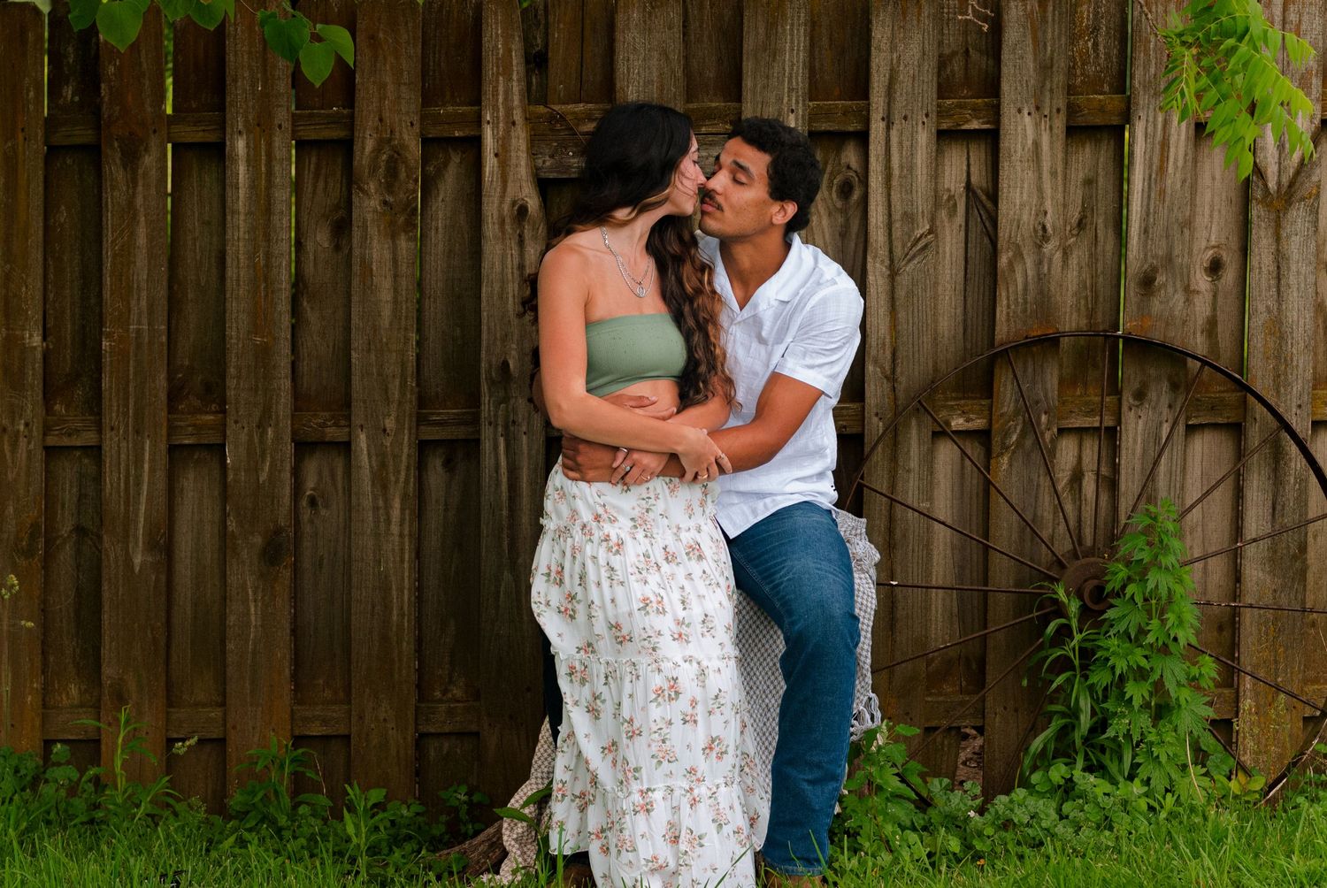 A couple sharing a romantic kiss in front of a rustic wooden fence with a vintage wagon wheel outdoors.