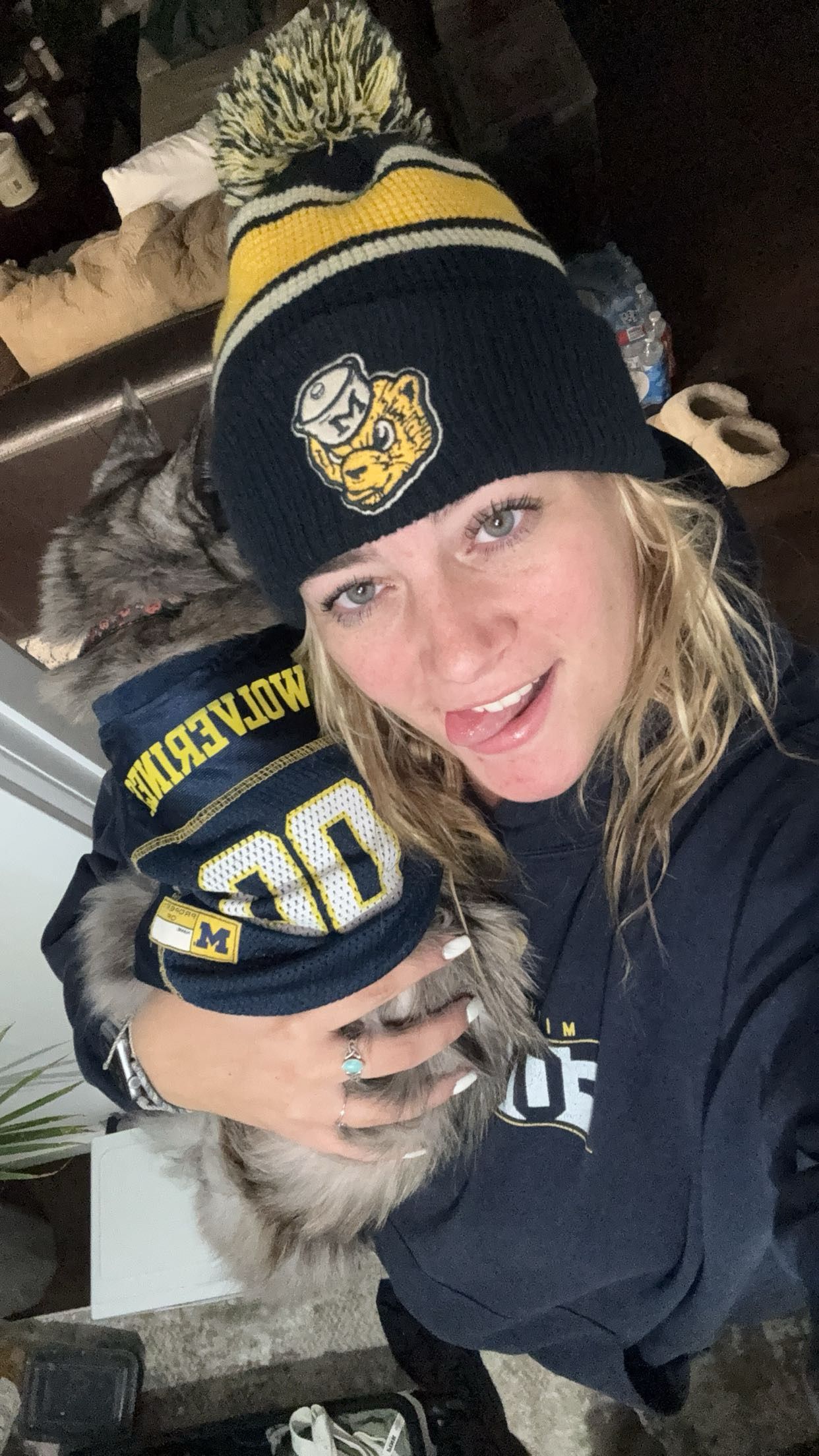 Woman wearing Michigan Wolverines beanie and sweatshirt holding a cat in a cozy indoor setting.