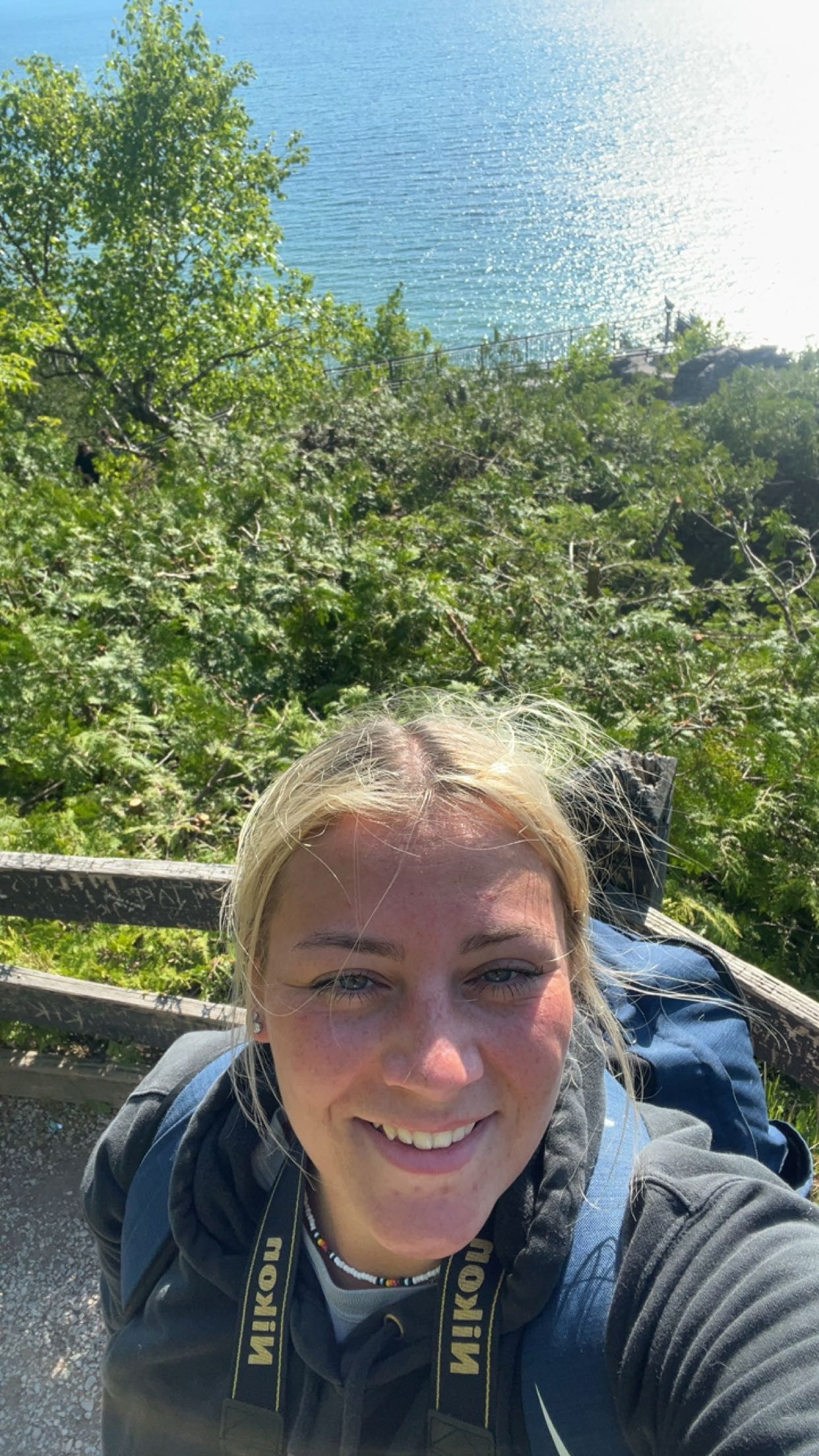 Smiling blonde woman takes a selfie while hiking with a Nikon camera strap, ocean view behind her.