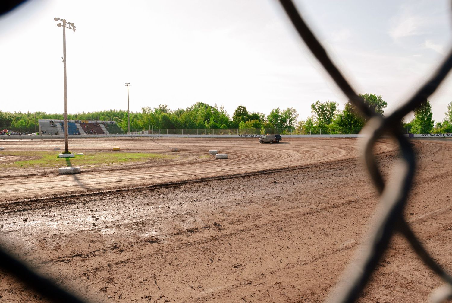 Empty dirt oval racing track viewed through chain-link fence on a cloudy day.