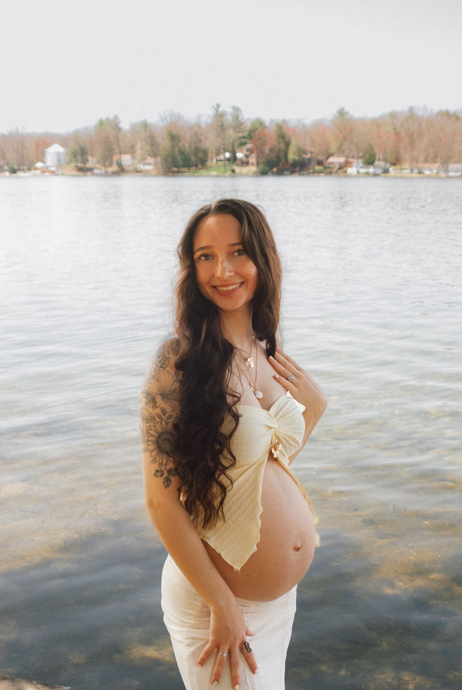 Happy pregnant woman with tattoos in cream outfit smiling by a lakeside setting.