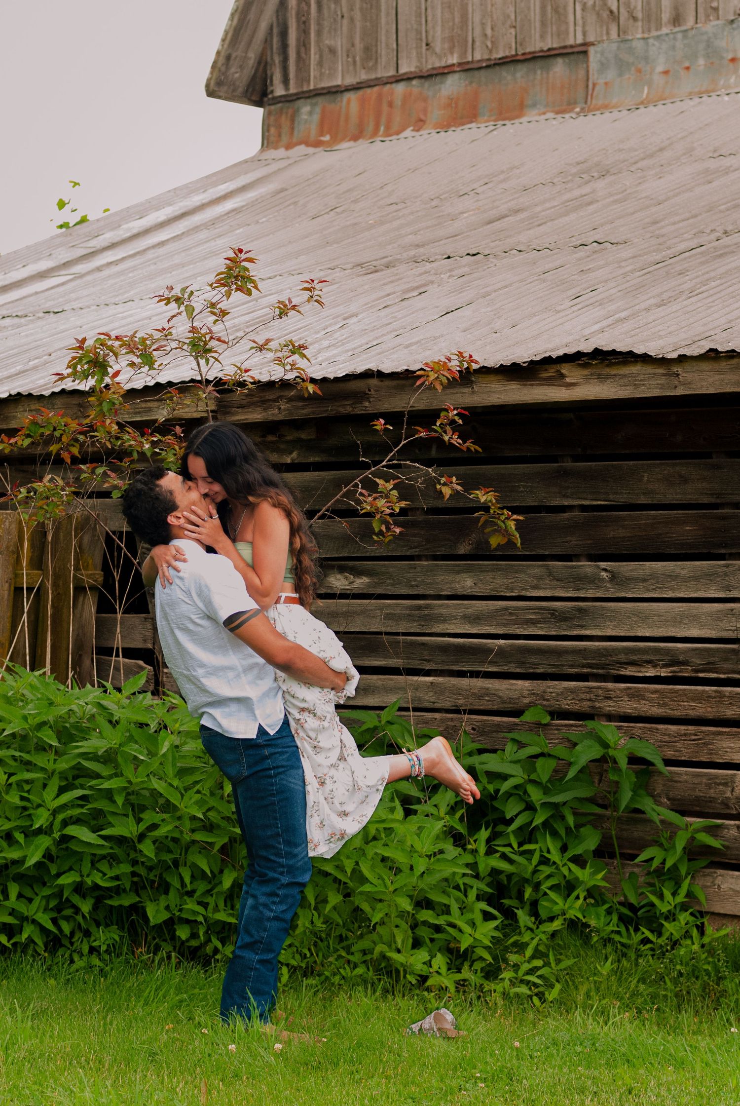 A joyful couple sharing a romantic lift and kiss in front of a rustic wooden barn outdoors.