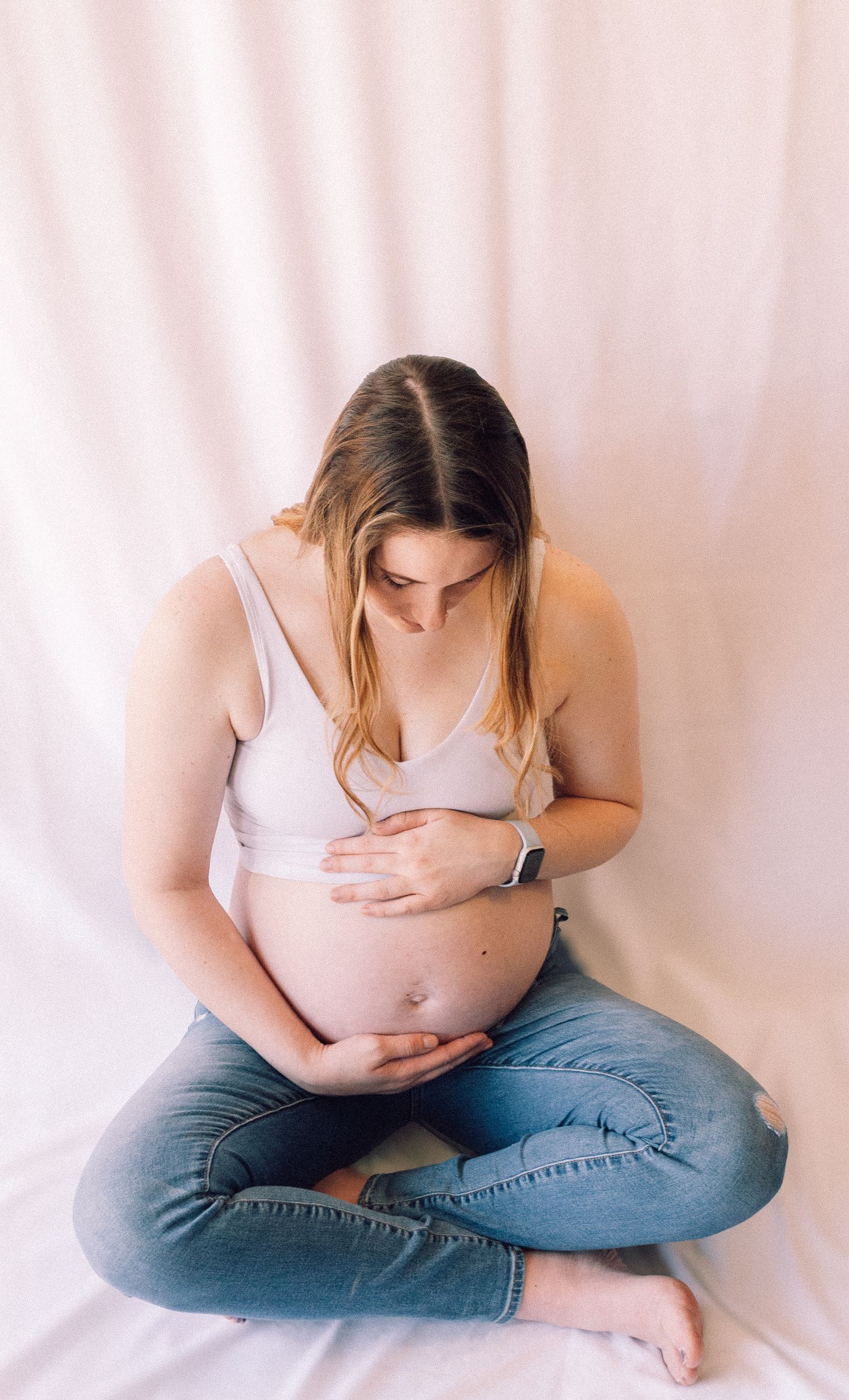 Pregnant woman in white bra top and jeans sitting cross-legged gazing down at her baby bump.