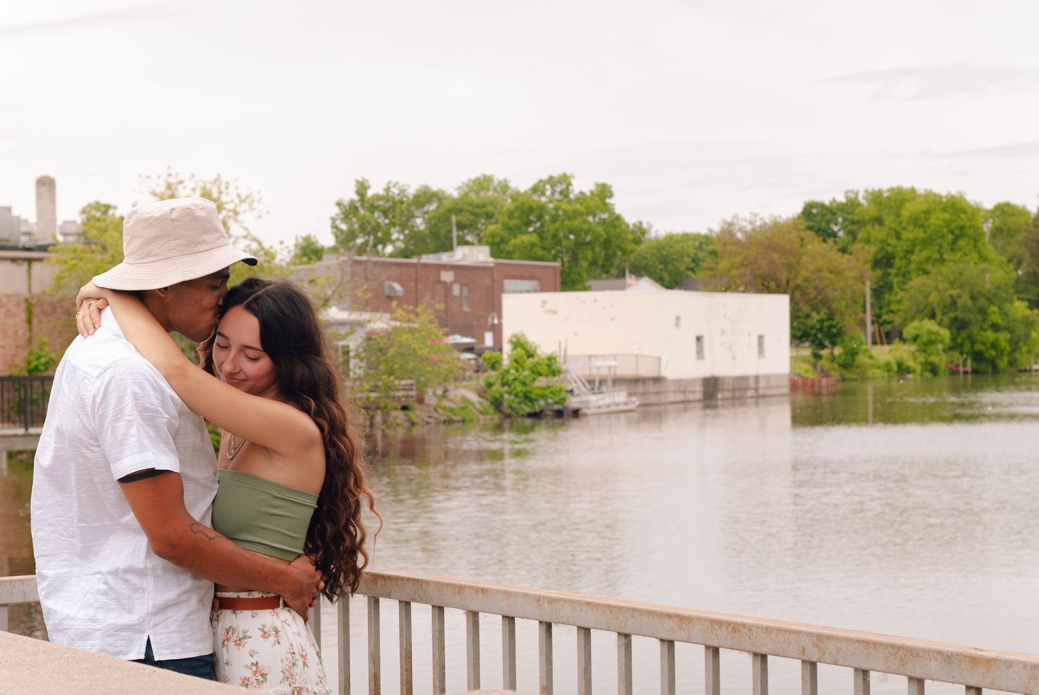 A couple embraces on a bridge overlooking a calm river with a historic town in the background.