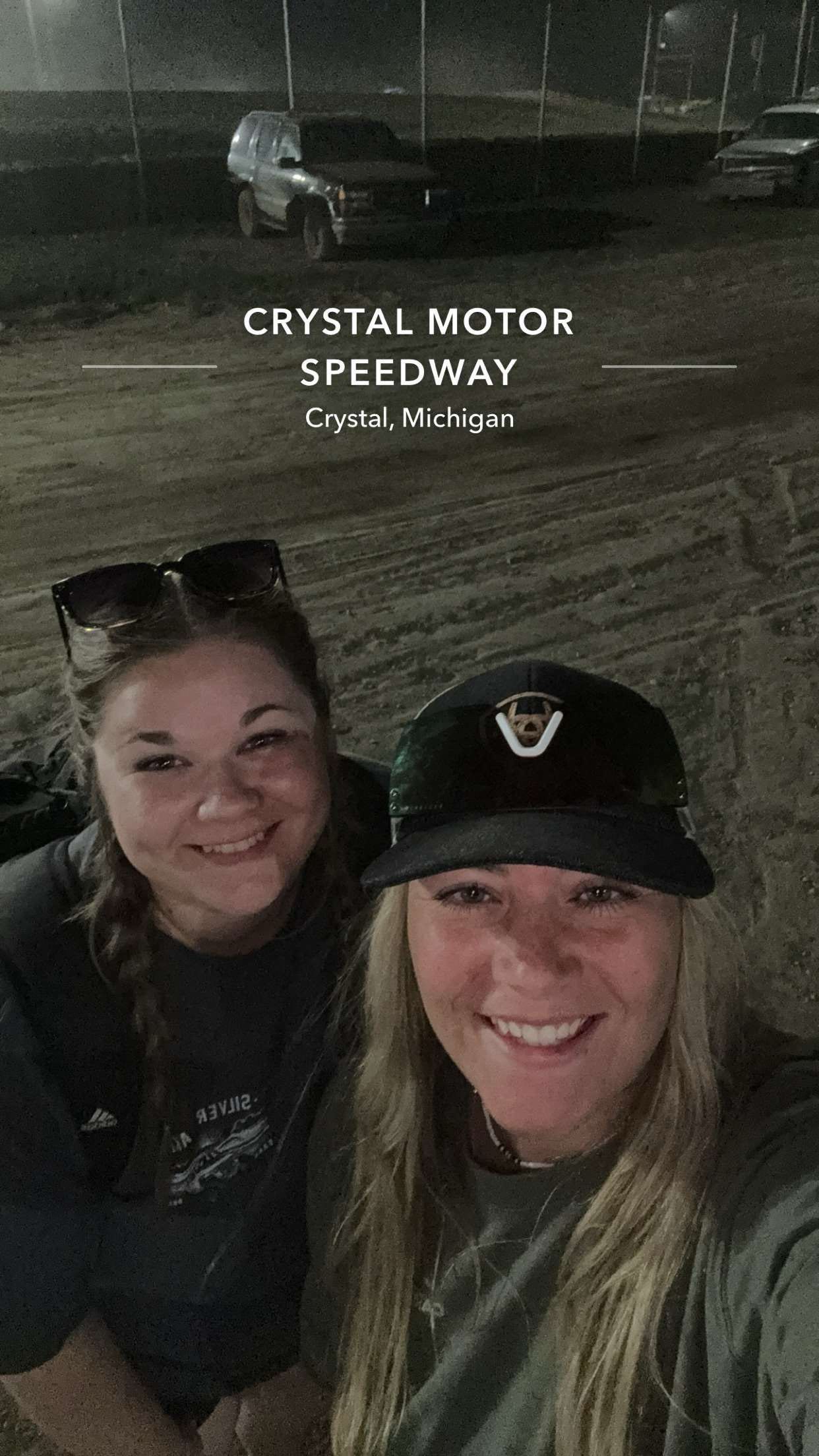 Two women smiling at Crystal Motor Speedway in Crystal, Michigan, with race cars on a dirt track visible in the background.