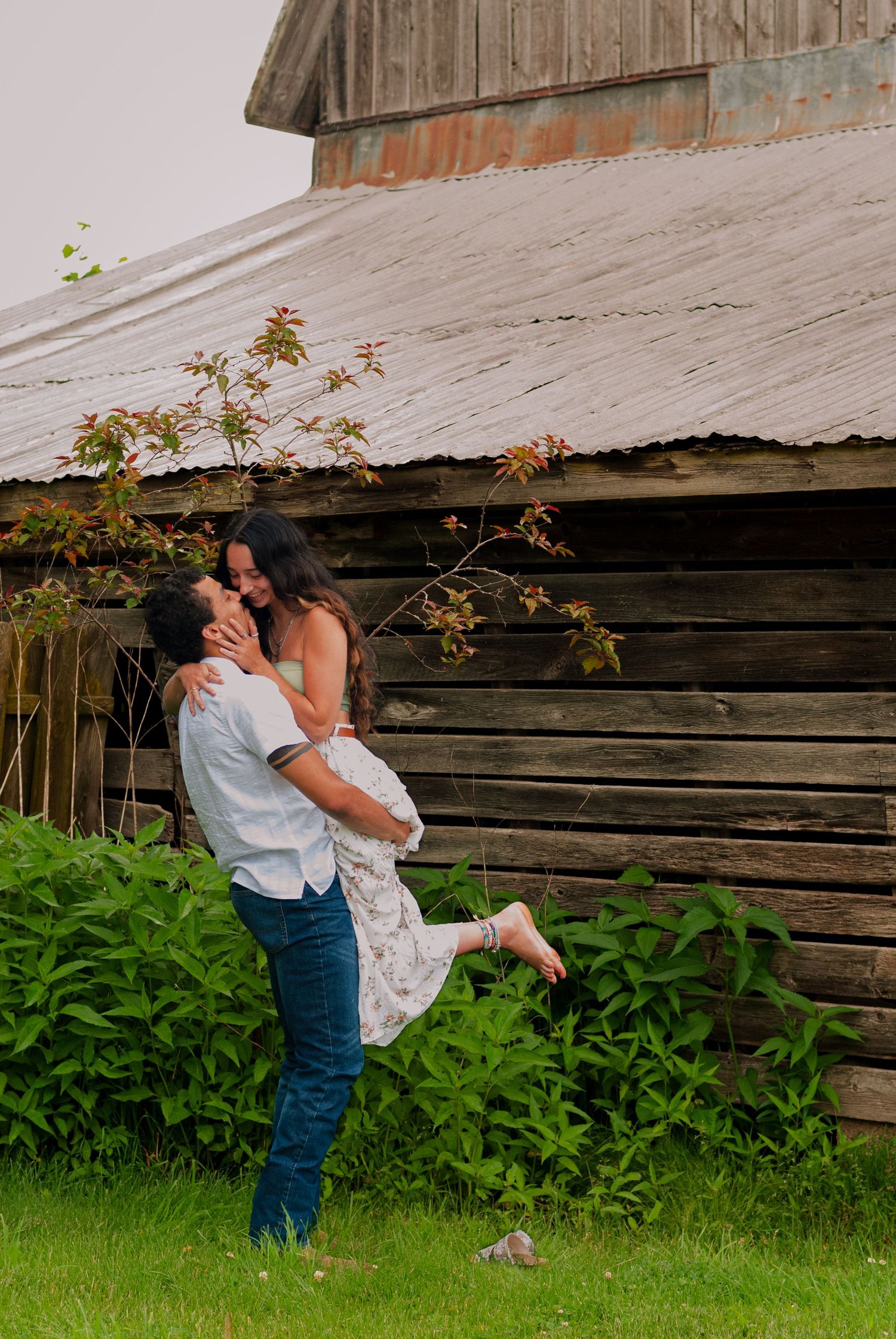 Couple embracing near rustic barn, man lifting woman in floral dress amid lush greenery.