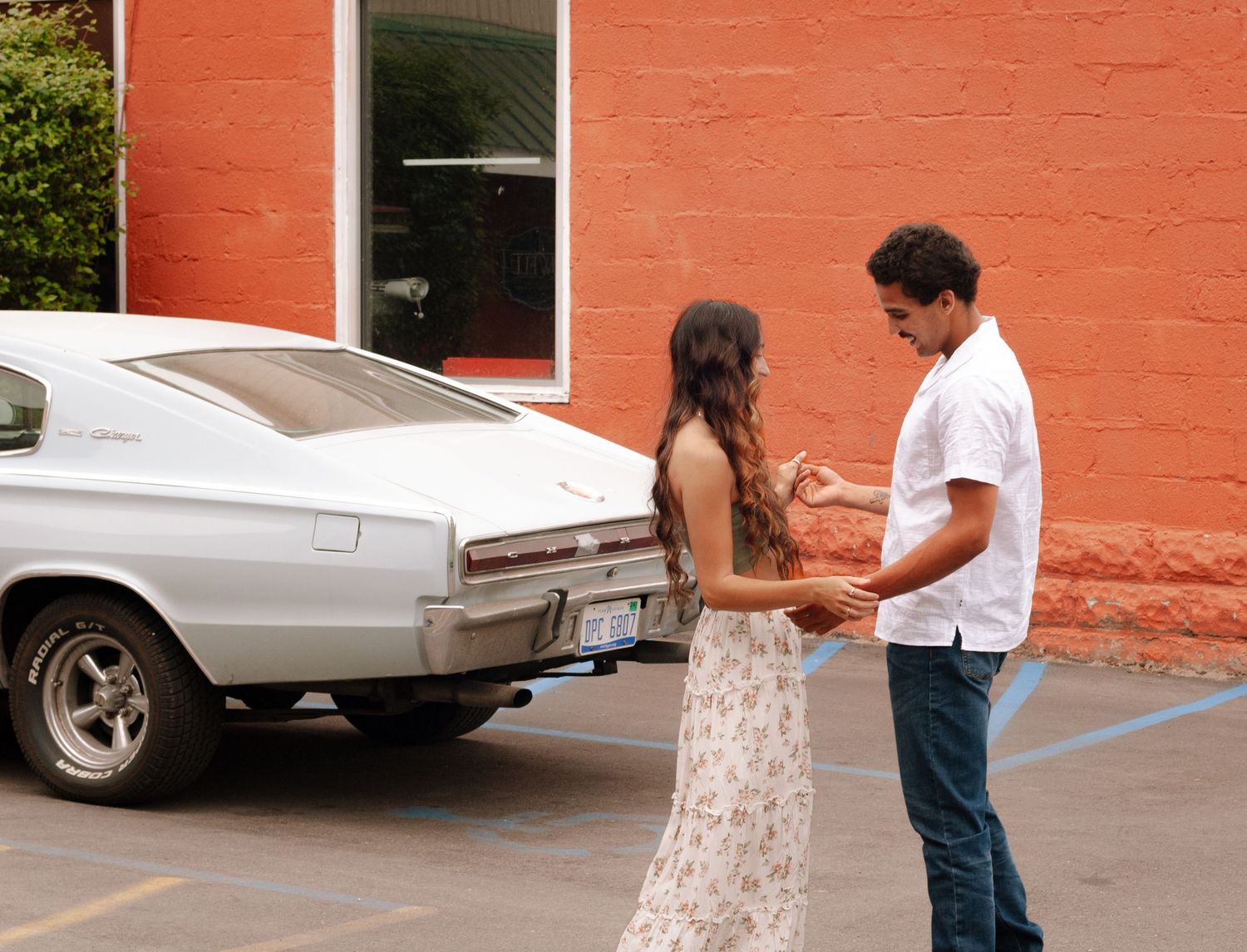 Young couple holding hands beside a classic white muscle car against an orange brick wall.