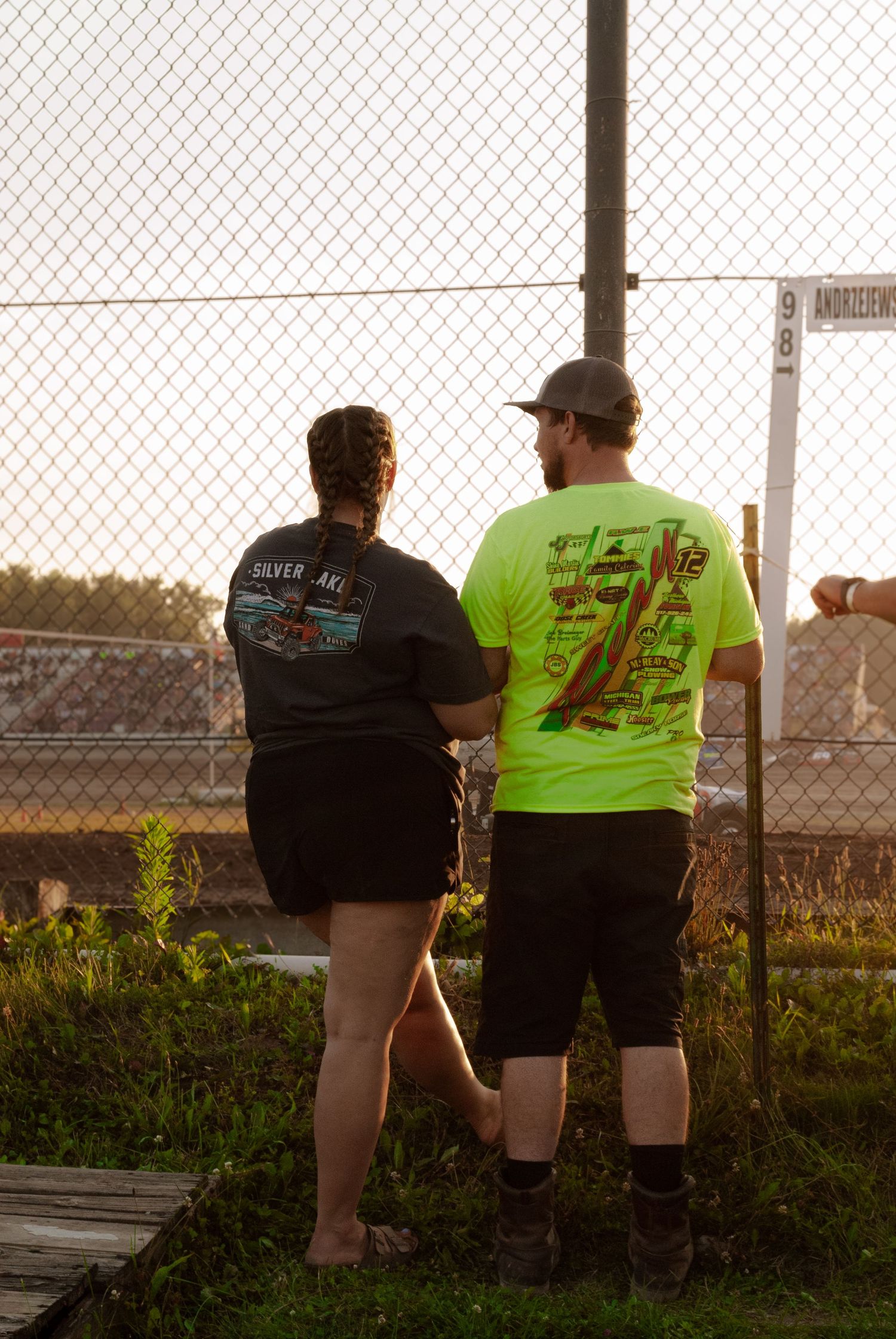 Couple watching dirt track racing through a chain-link fence at a local speedway at sunset.