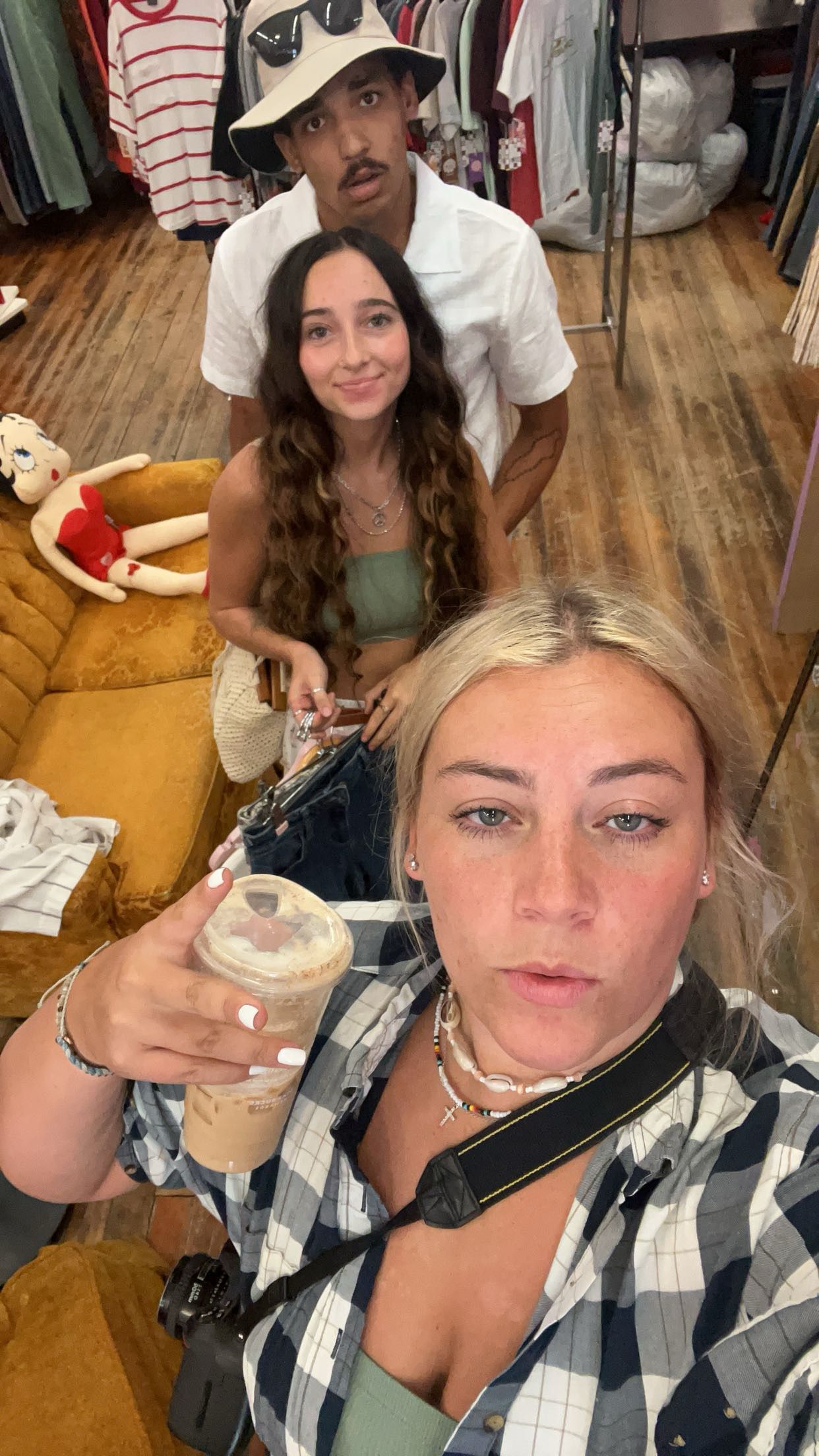Three friends take a selfie in a vintage clothing store, with one holding an iced coffee drink.
