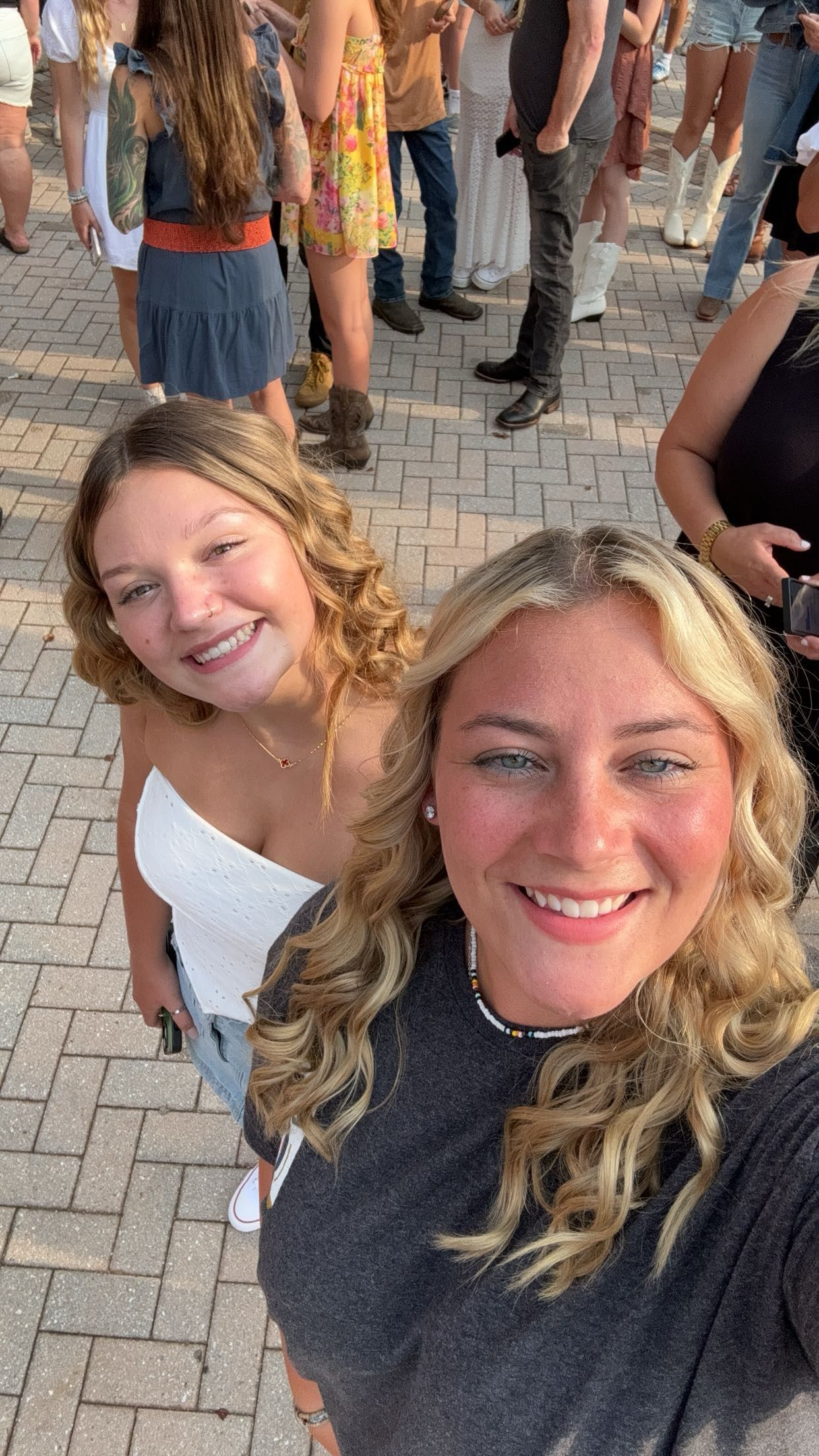 Two smiling blonde women with curly hair take a selfie outdoors at a crowded event on a brick plaza.