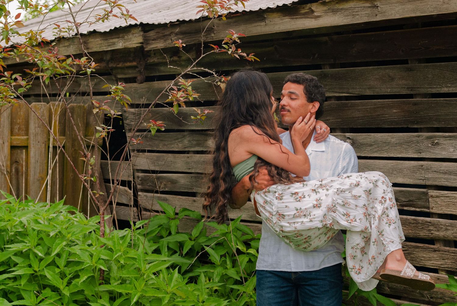 Romantic couple embracing near rustic wooden barn surrounded by lush green foliage.