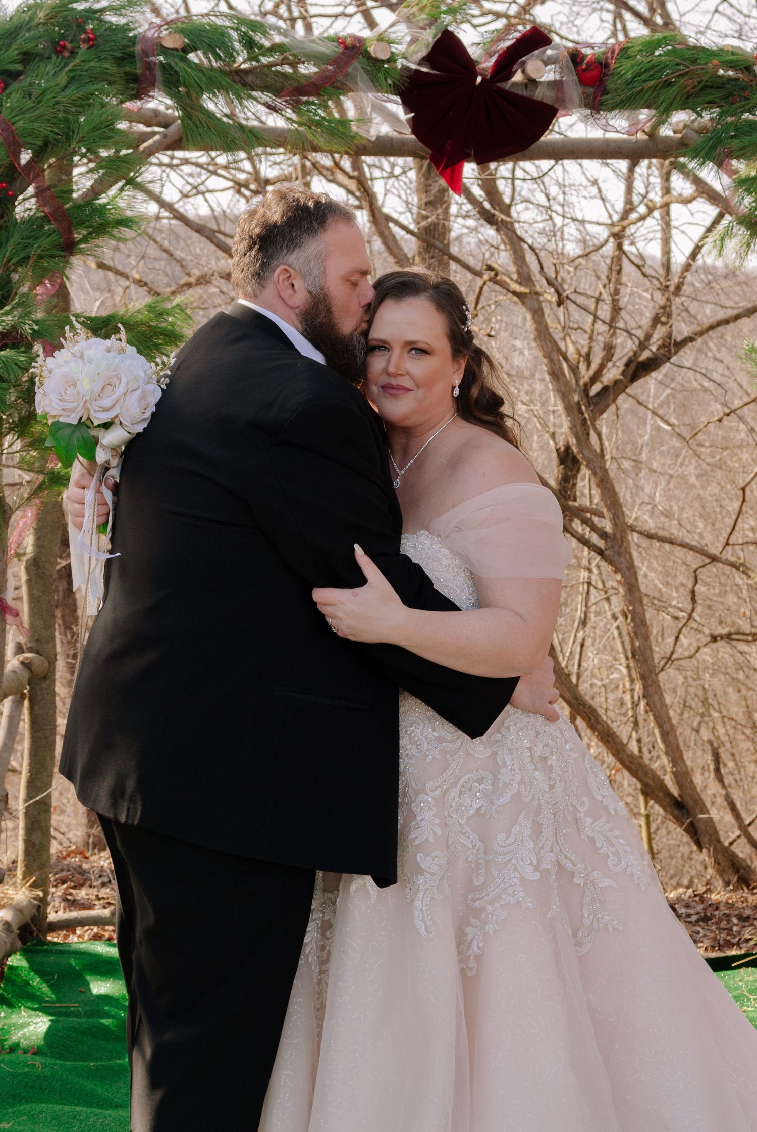 Groom kissing bride on cheek during romantic outdoor winter wedding ceremony under arch.