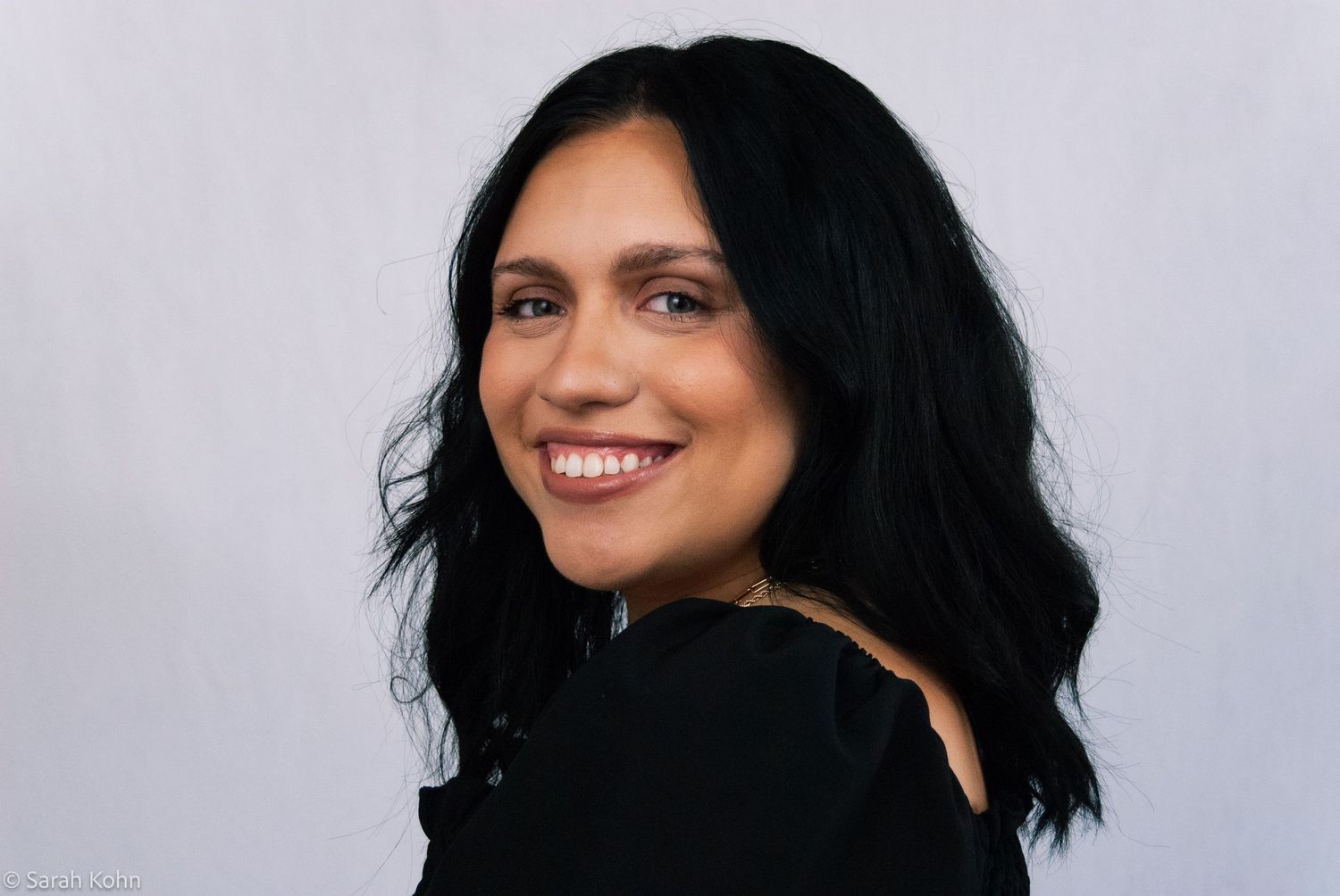 Smiling woman with black wavy hair wearing a black off-shoulder top against a light gray background.