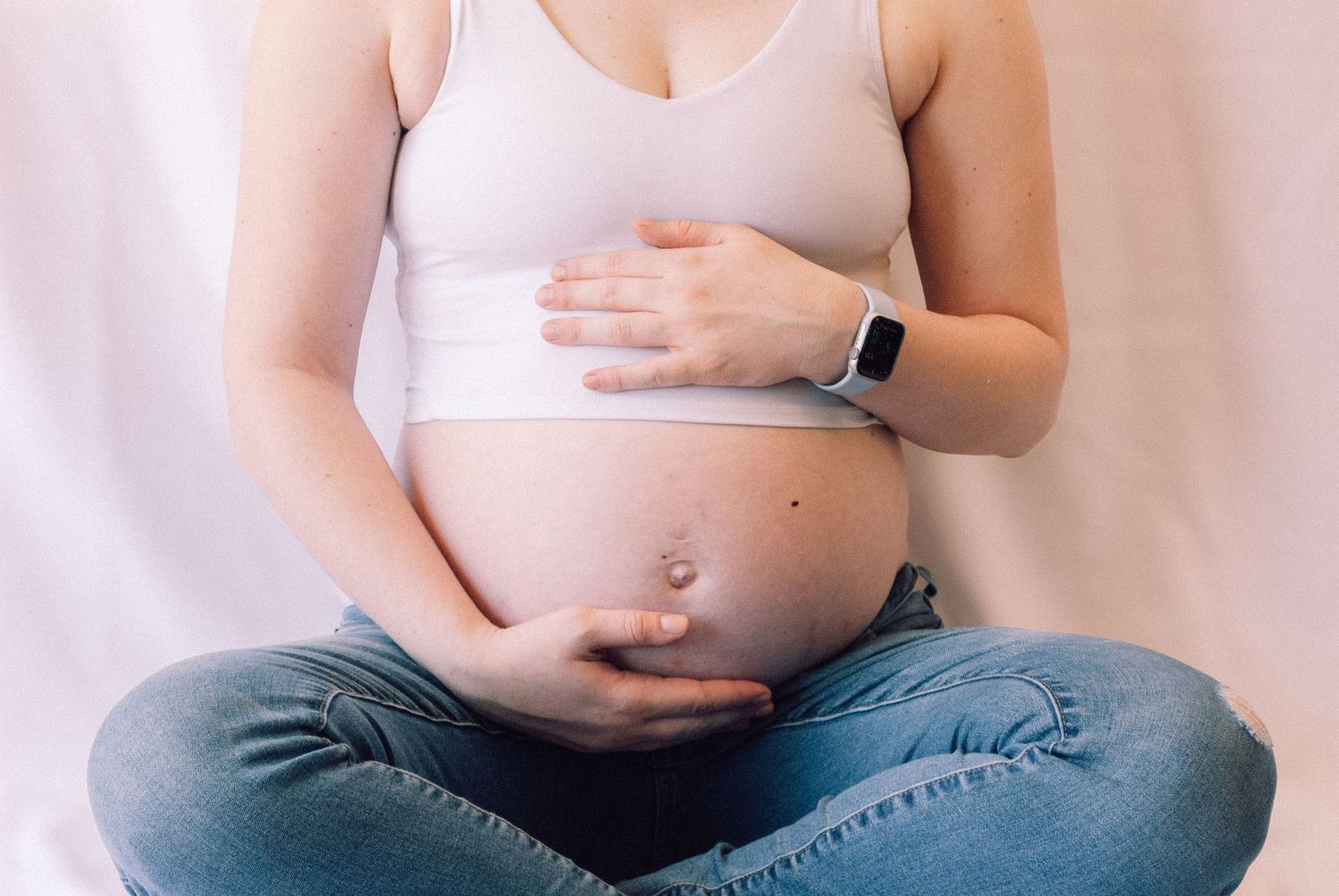 Close-up of pregnant woman in white bra top and jeans cradling her bare baby bump with both hands.